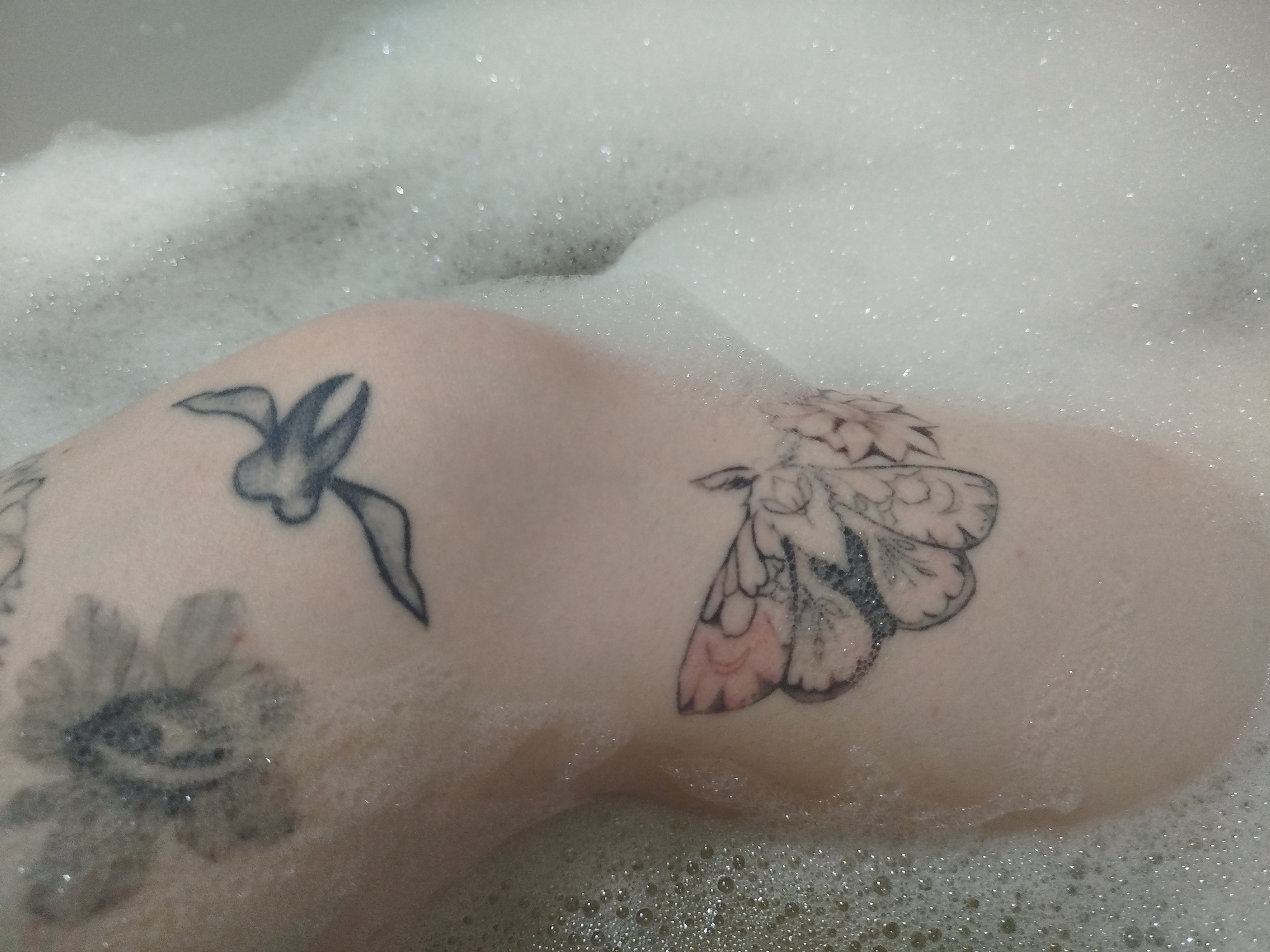 The image shows a person's knee and part of their leg submerged in a bubble bath. The skin is pale and has three visible tattoos: a flower, a stylized bird, and a moth. The bubbles are dense and cover most of the scene, creating a blurred and indistinct background. The location is not identifiable, and no information about the city or specific environment is available.