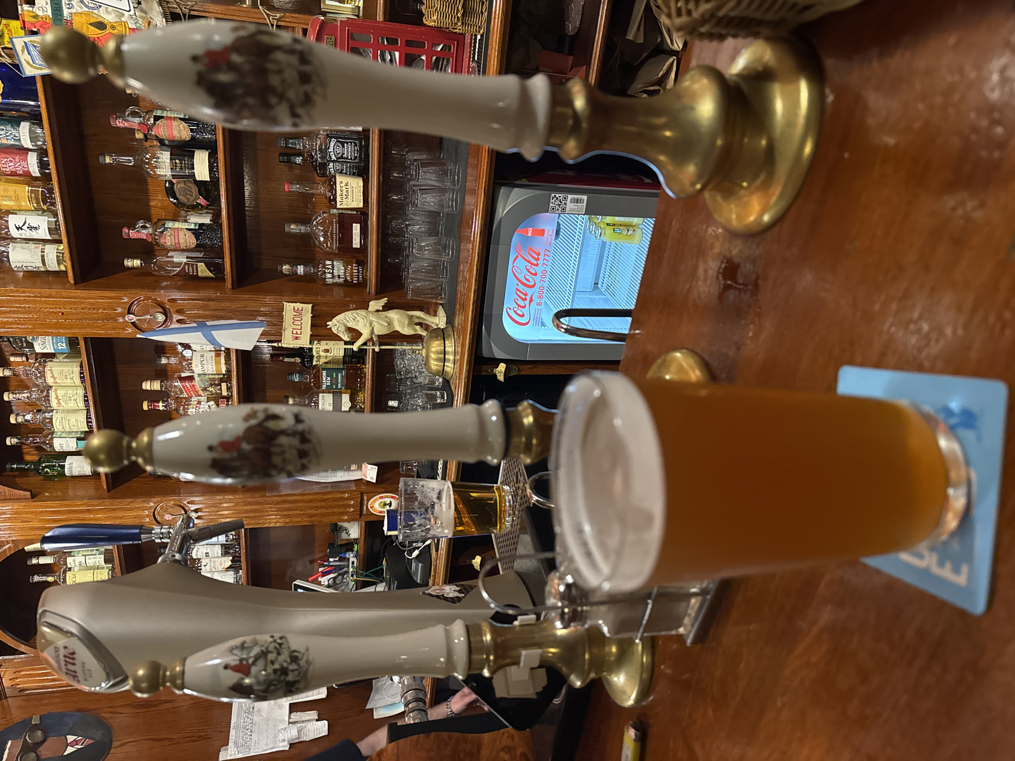 The image depicts a dimly lit bar interior, focused on a wooden bar counter with several beer taps and a half-full glass of amber liquid. The background reveals shelves stocked with various bottles of alcohol, a Coca-Cola mini-fridge, and decorative elements like a small white horse statue and a bell. The bar taps are ornate, with one featuring a depiction of a horse and rider. The overall atmosphere suggests a traditional pub setting. There is no information available to determine the location of Saint Petersburg, Russia.