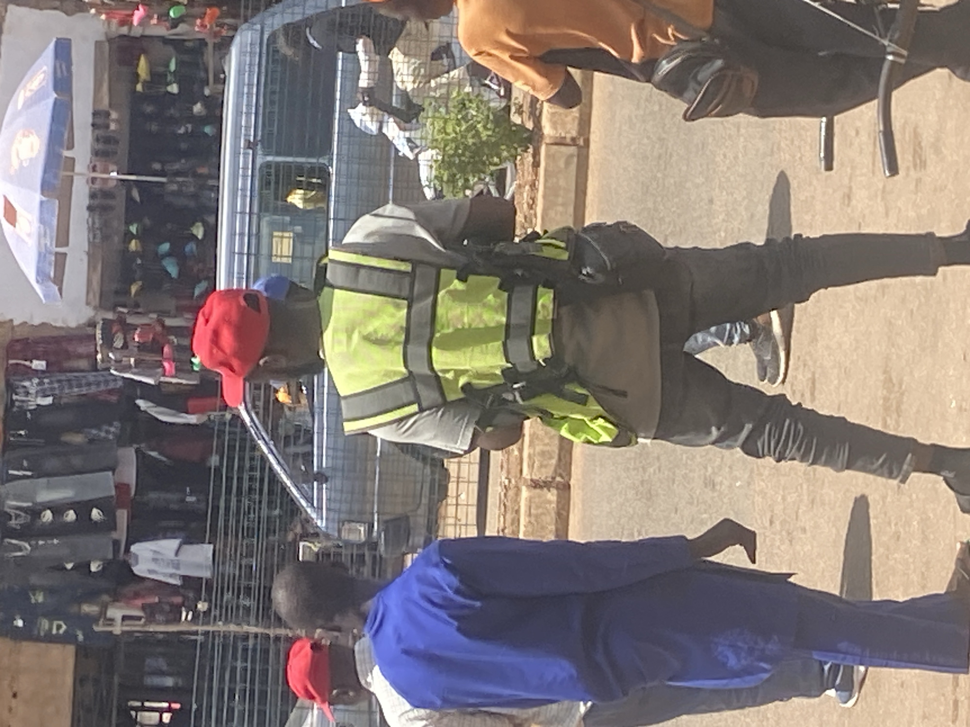 The image depicts a street scene in Bukuru, Nigeria, likely a market area. In the foreground, three individuals are visible from the waist down, with a fourth person partially visible on the far right. The central figure, wearing a red cap and a high-visibility neon yellow vest over a grey shirt, appears to be a security guard or traffic personnel, as indicated by the vest and the tactical pouches attached to his belt. He is wearing dark pants. To his left, a person in a bright blue robe and white shoes is facing away from the camera. Another individual, also wearing a red cap, is mostly obscured by the person in blue. On the right, a person in an orange shirt and black pants is visible from the side. The background shows a row of stalls displaying clothing and other goods, with an open-fronted shop displaying shoes under a blue umbrella. A light-colored van is parked behind the people in the foreground, suggesting an urban street or market setting. The overall impression is one of a bustling commercial area in Nigeria.