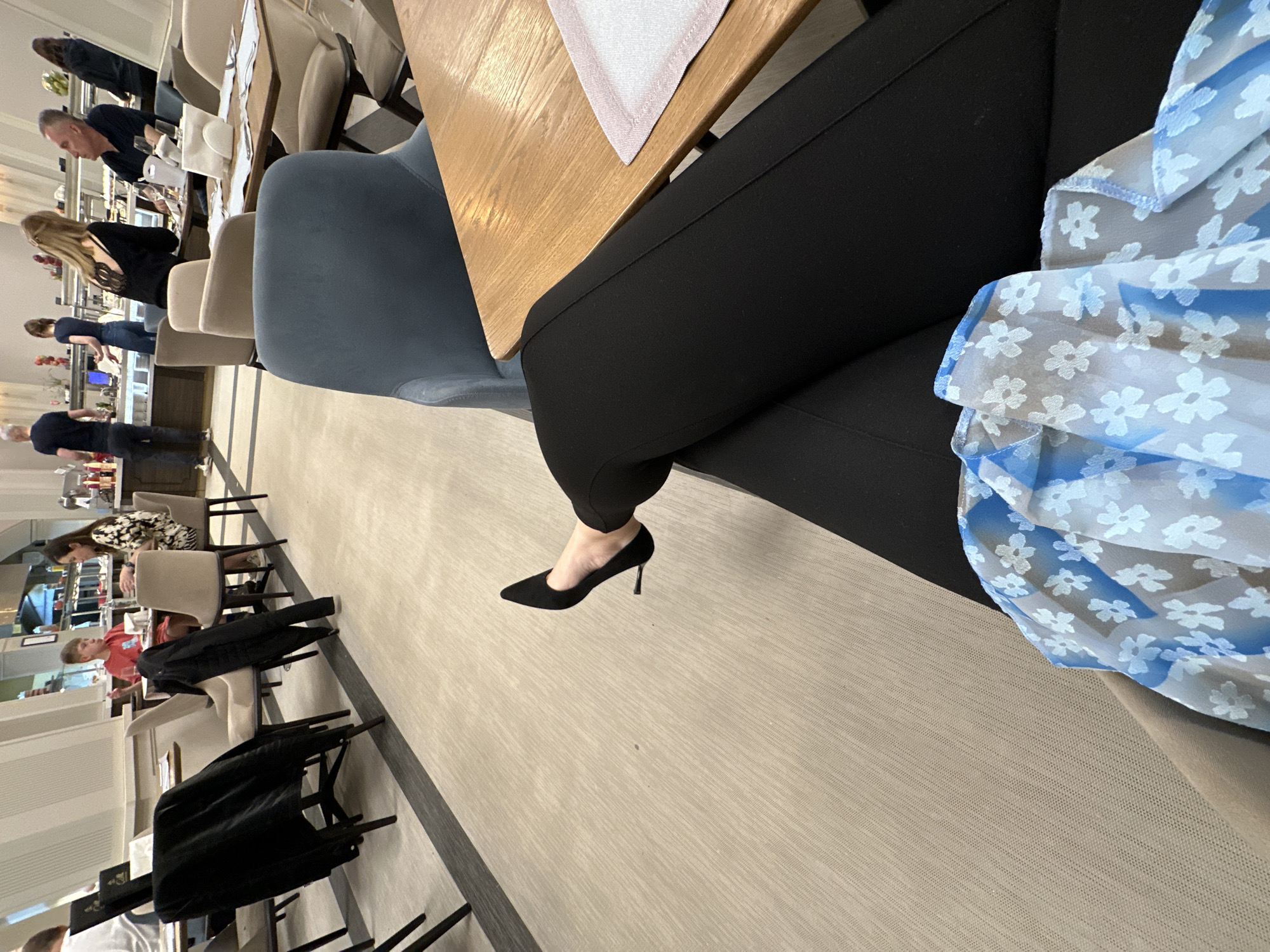 The image shows a person seated at a wooden table in a dining area, possibly a restaurant. The person is wearing black pants and a blue floral-patterned garment. Their foot is visible, clad in a black high-heeled shoe. Other people are seated at tables in the background, and there is a buffet setup along the wall. The location is suggested to be in the city of Kazanlak, Bulgaria.