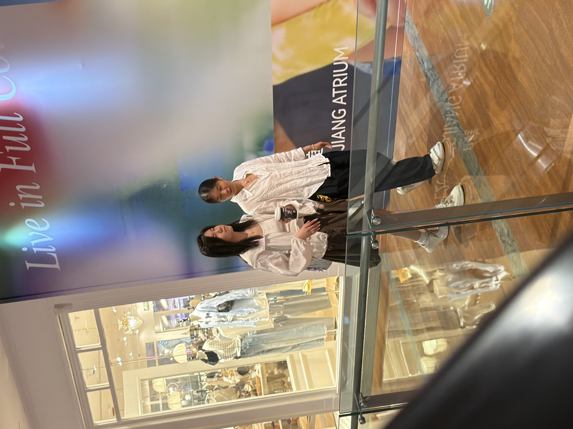The image shows two young women indoors, with a large poster behind them. The woman on the right is wearing a white collared shirt and black pants. She is holding a cup in her hand and appears to be looking at her phone. The woman on the left is also wearing a white collared shirt, and she has her arm around the other woman. They are both looking down and slightly away from the camera. The poster behind them has text that says "JIANG ATRIUM" and "Live in Full Co". The floor appears to be made of polished wood. In the foreground, a glass railing is visible. Below the railing is a store window displaying clothing on mannequins and shelves. The lighting suggests it is daytime, but the specific time of day is not discernible. The overall scene appears to be in a shopping mall or a similar commercial space.