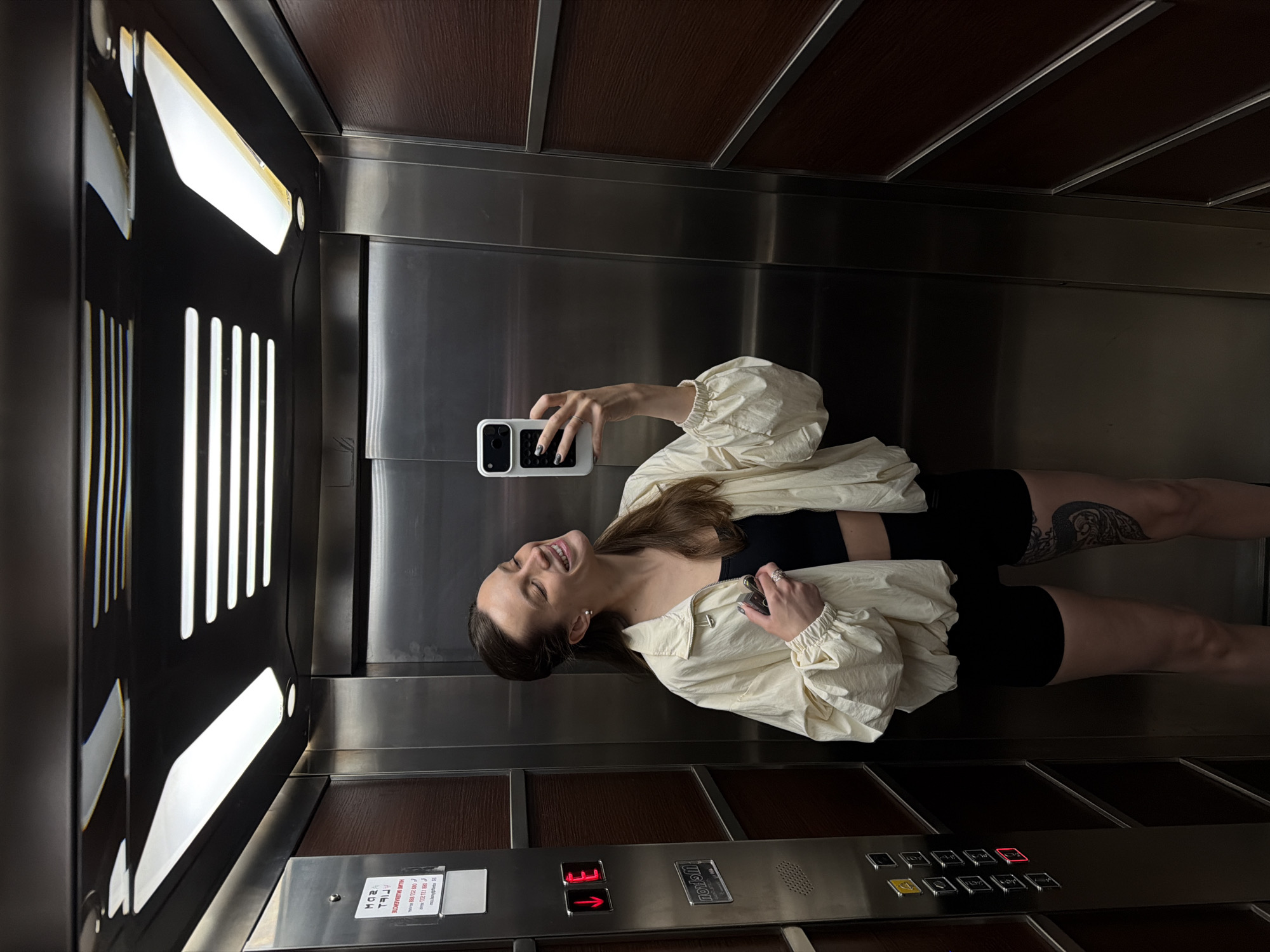 A woman is taking a selfie in an elevator. She is wearing a black top and shorts with a cream-colored jacket. The elevator has wooden and steel paneling. The button panel shows the number 3 illuminated in red. There is a small sign near the bottom of the panel. The specific location in Budva, Montenegro cannot be determined from the visual information.