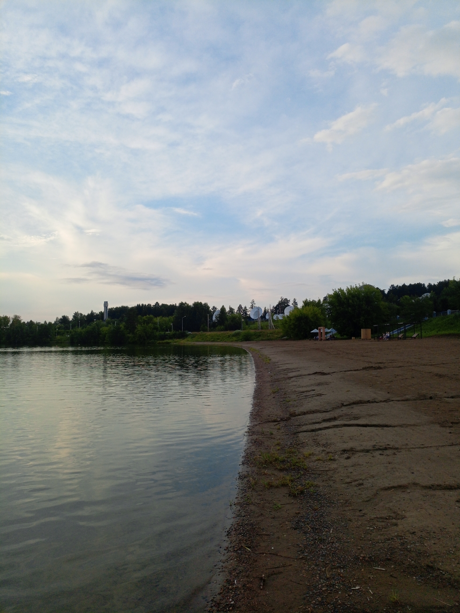 This image depicts a body of water with a sandy shore in Zheleznogorsk, Russia, likely a recreational area. The water's surface is calm, showing ripples and reflections of the sky and surrounding trees. The shore is composed of sand and small pebbles, with some sparse vegetation growing along its edge. In the background, a dense line of trees forms a natural backdrop, with some buildings and structures visible among them. One prominent structure is a tall, slender tower with a square base. Further along the shore, several large, parabolic satellite dishes are visible on raised platforms. There are a few beach umbrellas and some wooden structures on the sand, suggesting a public beach or park area. The sky is partly cloudy with a mixture of white and blue tones, indicating it is either early morning or late afternoon. No individuals are clearly visible in the image, but the presence of beach amenities implies potential human activity in the area. The overall scene is tranquil and naturalistic, with a touch of technological infrastructure in the form of the satellite dishes.