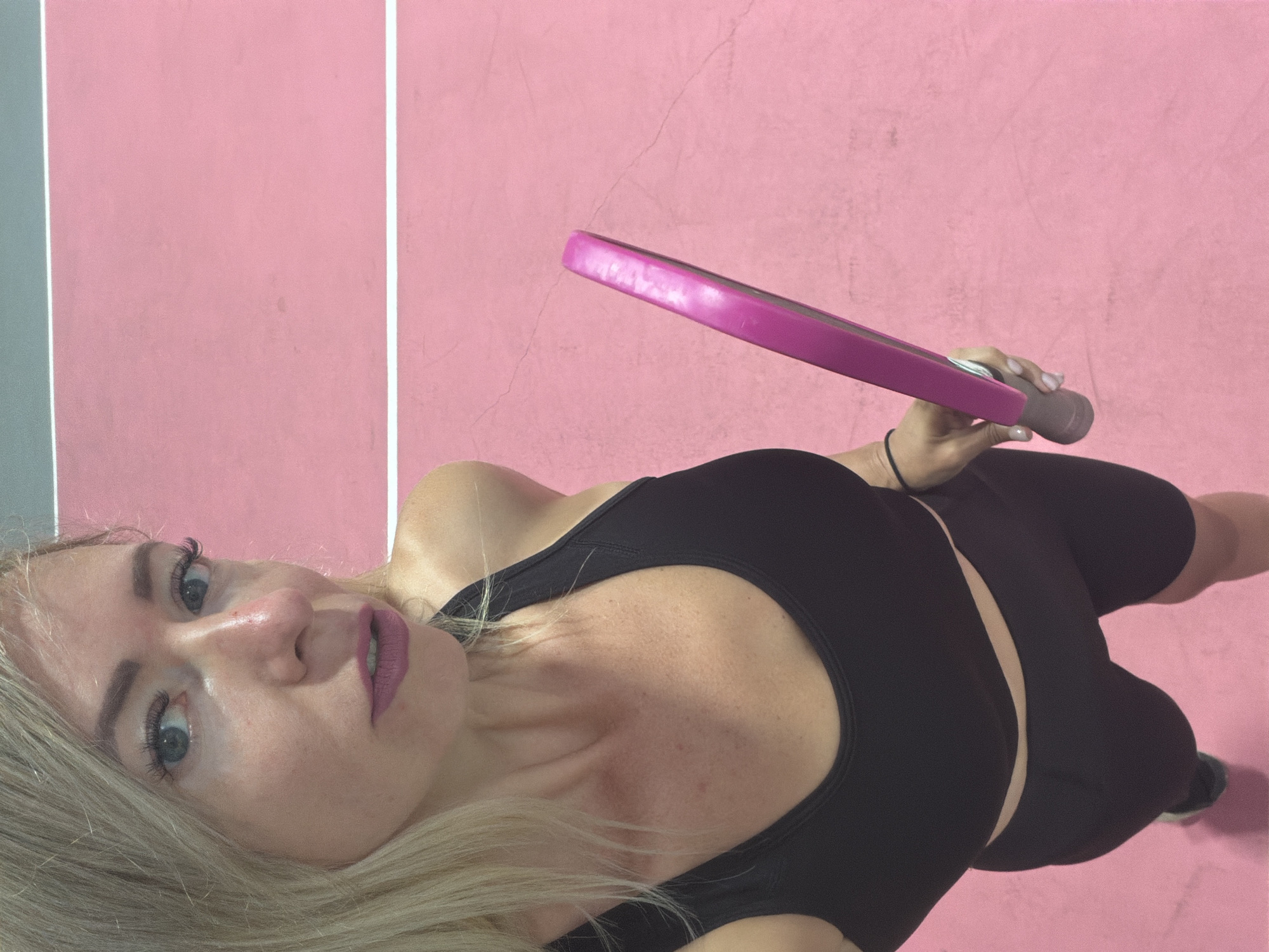 A woman with blonde hair and blue eyes is taking a selfie on a pink court. She is wearing a black sports bra and black bike shorts. She is holding a pink paddle in her right hand. The court has white lines on it.