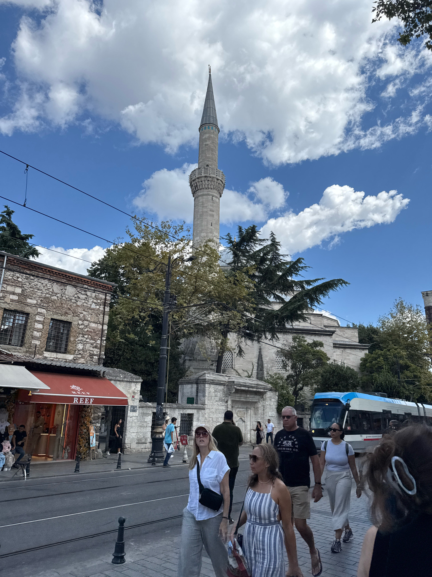 In Istanbul, Turkey, a bustling street scene unfolds with a prominent minaret of a mosque rising against a bright blue sky dotted with white clouds. In the foreground, several individuals are walking on a sidewalk adjacent to a street with tram tracks.
On the left side of the image, a stone building with arched windows is visible, with a shopfront featuring a red awning and the "REEF" logo. People are seen browsing and walking past the shop.
As the scene moves to the right, more pedestrians are captured in motion. A woman in a white shirt and gray pants with a black crossbody bag walks towards the viewer. Beside her, a man in a black t-shirt and khaki shorts walks with a woman in a white top and light-colored pants. Another woman in a striped dress is walking slightly ahead of them.
In the background, a modern tram with blue and white accents travels along the tracks. The architecture of the mosque, with its intricate details and dome, is visible behind the trees and streetlights. The overall atmosphere is that of a lively urban environment on a clear day, with a mix of historical and modern elements.
