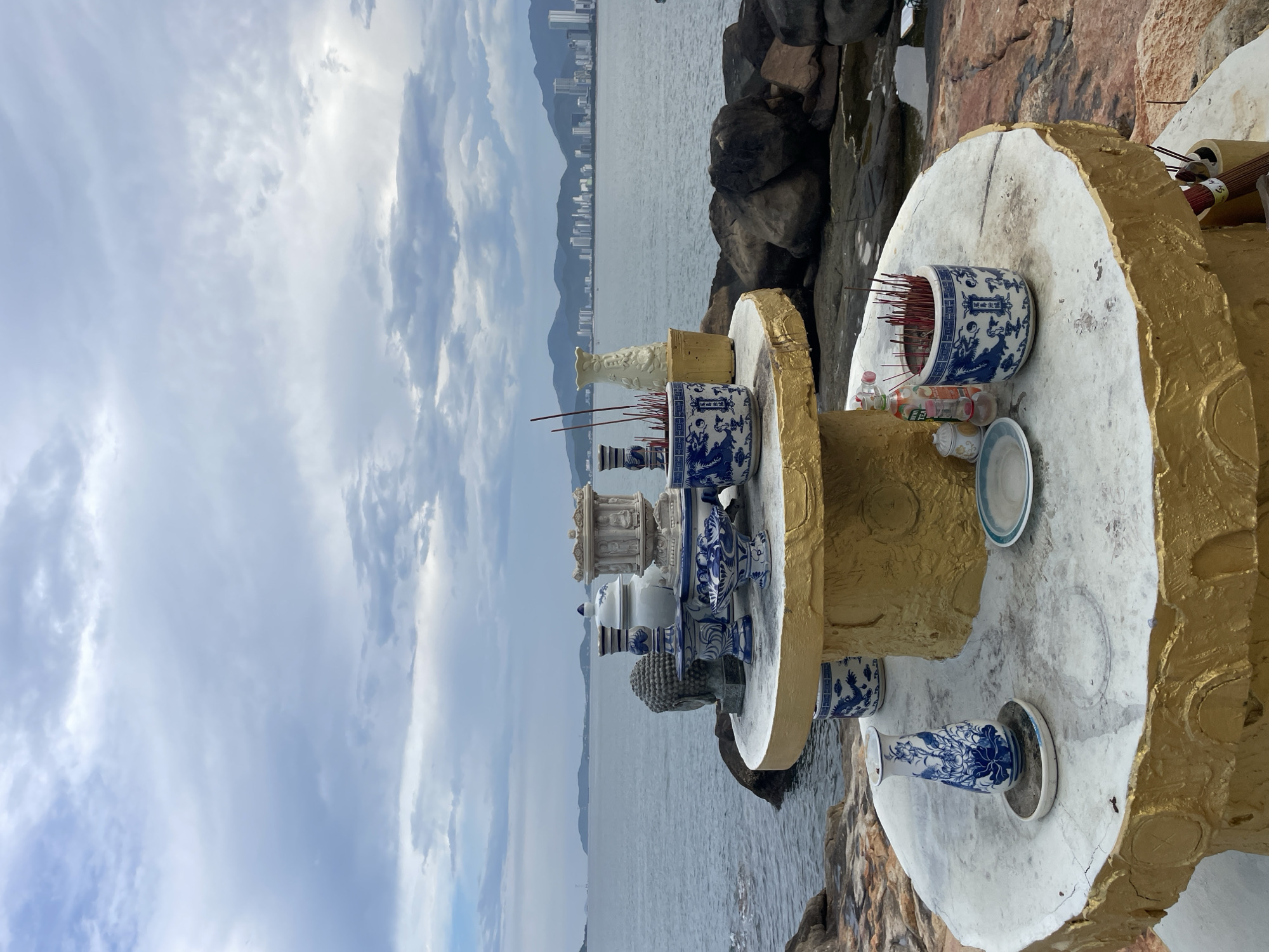 This media file captures an outdoor shrine situated on a rocky coastline, likely in Nha Trang, Vietnam. The shrine is adorned with several ceramic items, predominantly blue and white, featuring intricate dragon motifs and floral patterns. Among the objects are incense burners filled with red incense sticks, a vase, and what appears to be a Buddha head sculpture. The shrine itself has a weathered, white surface with gold trim, suggesting it is exposed to the elements.
The setting is a coastal landscape with a body of water stretching towards a distant city skyline and mountainous terrain. The sky is overcast with dramatic clouds, indicating a possibly moody or stormy weather condition. The water is a calm grey, reflecting the cloudy sky. The rocks surrounding the shrine are rough and varied in color, from dark grey to reddish-brown.
There is no clear indication of people or specific activities taking place within the frame, beyond the presence of the shrine itself, which implies a devotional or memorial purpose. The overall impression is one of a serene yet somewhat somber atmosphere, with the natural elements of the sea and sky dominating the background. The time of day appears to be daytime, given the ambient light, but the overcast conditions make it difficult to ascertain the exact time. No visible text is present in the image.