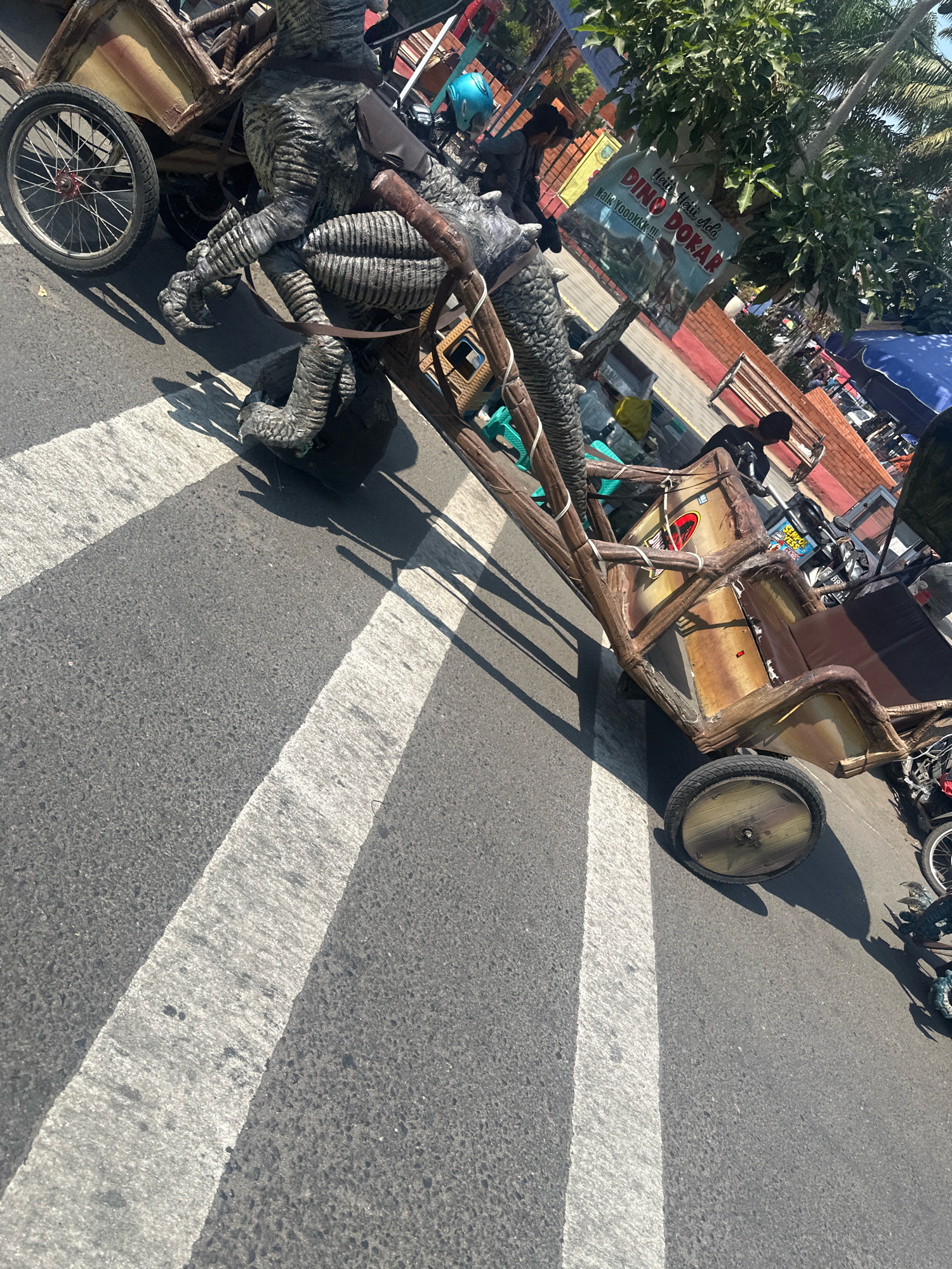 An elaborate, custom-built, two-wheeled cart, resembling a dinosaur-themed float, is positioned on an asphalt road marked with white pedestrian crossing lines. The cart features a faux-wood structure, and a large, grey, reptilian-like figure with prominent claws and textured skin is the main subject, appearing to be part of the cart's design or a driver. The figure is partially obscured by the cart's structure. In the background, a sign with Indonesian text, including "Dino Dokar," is visible, suggesting a local attraction or business. Several people are present, some appearing to be operating or interacting with the cart, while others are in the background, possibly pedestrians or vendors. The environment suggests a public street or marketplace in Mojokerto, Indonesia, under bright daylight conditions, as indicated by strong shadows cast by the objects. The overall scene depicts a colorful and somewhat whimsical street display.