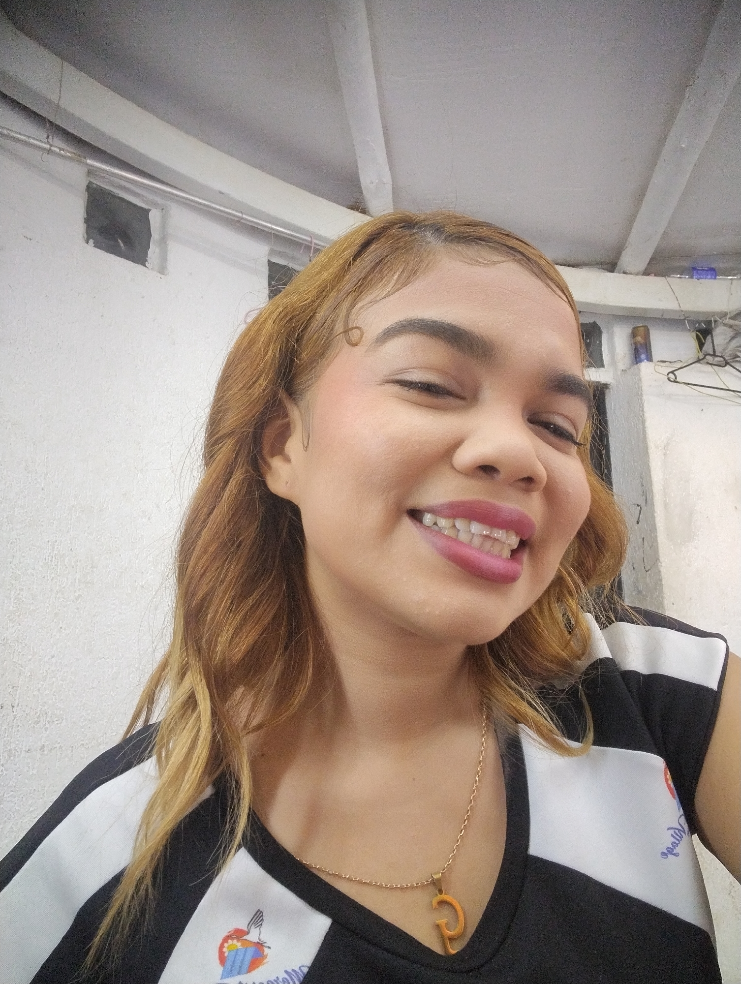 A woman is smiling and looking slightly to the left. She is wearing a black and white top with a logo of a bird over a map of Venezuela and writing in blue, and a gold necklace with a letter "p" pendant. She has light brown hair and is wearing pink lipstick and blush. The background shows a white wall with exposed pipes and possibly concrete or plaster ceiling with some hanging objects. Given the logo on her shirt featuring a map of Venezuela, and the instruction to include the city of Porlamar, Venezuela, it is reasonable to state that the image was likely taken in Porlamar, Venezuela, though the specific location within the city cannot be determined from the provided information.