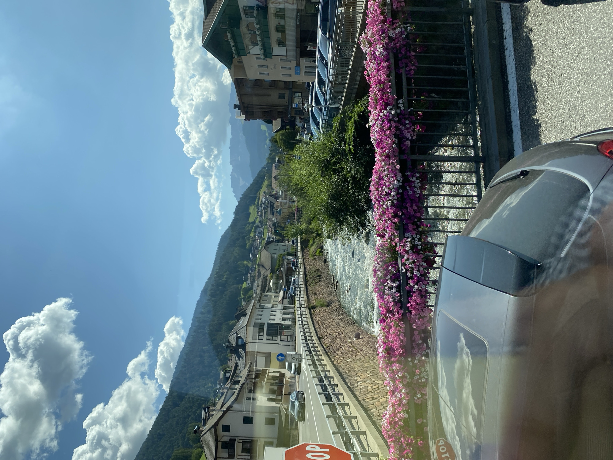 The image depicts a sunlit street scene in Ortisei, Italy, with a clear blue sky dotted with fluffy white clouds. A narrow river flows alongside the road, bordered by a stone wall. A metal railing above the wall is adorned with an abundance of pink and white flowers. Several buildings, typical of alpine architecture with tiled roofs, line the opposite side of the street. Cars are parked along the roadside, and a few are in motion on the street. In the foreground, the roof of a car with a sunroof is visible. The overall impression is one of a picturesque town nestled within a mountainous landscape.