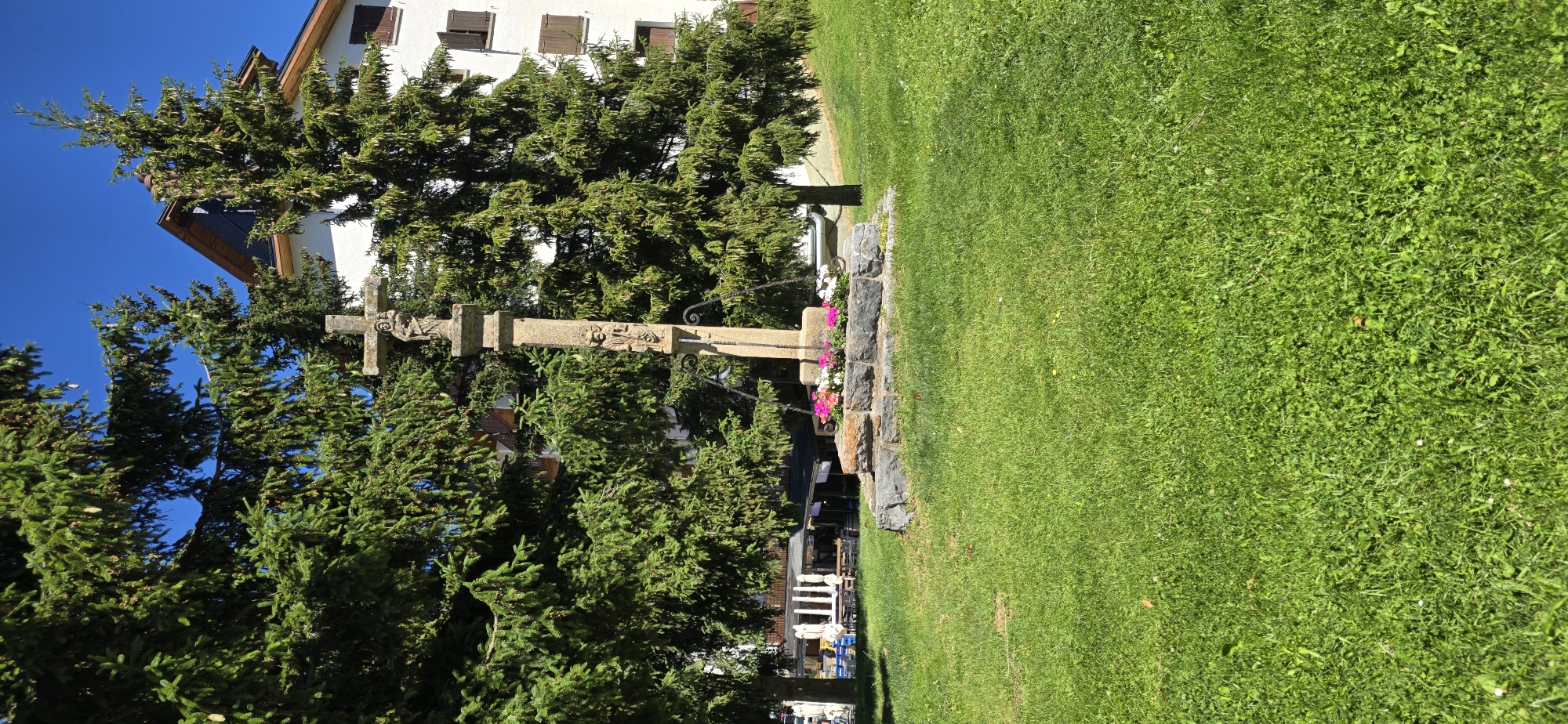 This media file depicts a stone wayside cross situated in a grassy area adjacent to a building, with numerous evergreen trees framing the scene. The cross itself is ornate, featuring carvings on its upright and horizontal beams, and appears weathered. It is positioned on a stone base adorned with a small arrangement of colorful flowers, predominantly pink and white.
In the background, a white building with brown shutters and a dark-tiled roof is visible, suggesting a residential or institutional structure. The surrounding trees are dense and green, creating a natural, somewhat enclosed atmosphere around the cross. Beyond the trees, hints of other buildings and infrastructure, including tables and chairs, suggest the presence of a village or town.
The activity or event taking place is not explicitly shown, but the setting implies a peaceful outdoor environment, possibly a memorial site or a landmark within a community. The bright sunlight and clear blue sky indicate a pleasant, sunny day. The overall mood is serene and perhaps a little solemn due to the presence of the cross. There is no visible text in the image. The location context suggests Sallent de Gállego, Spain.