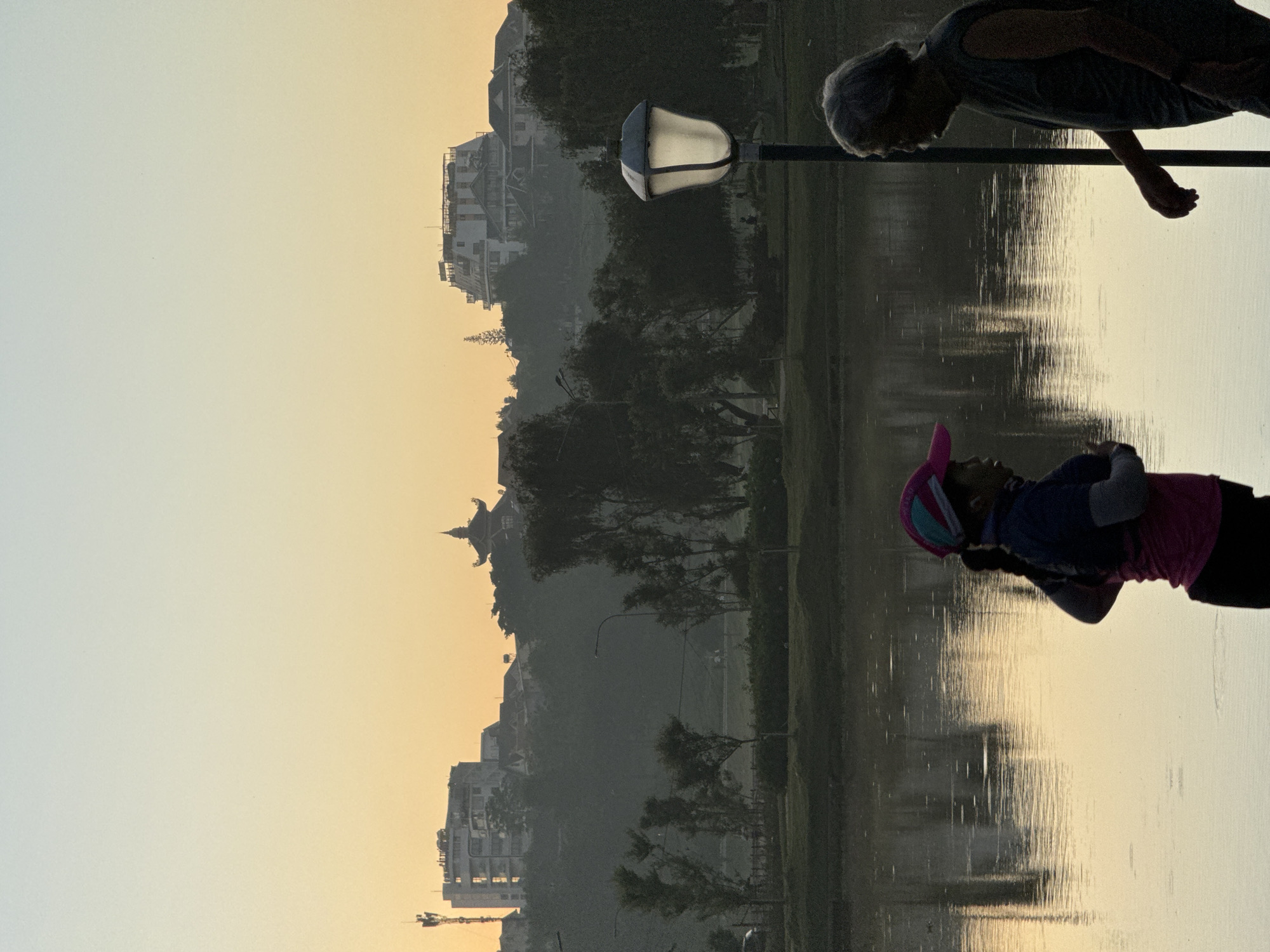 The image depicts a serene morning scene by Xuan Huong Lake in Da Lat, Vietnam. The sky is a gradient of soft orange and pale yellow, indicative of sunrise. Two figures are in the foreground, silhouetted against the water. On the left, a person wearing a pink cap and a blue and pink athletic outfit appears to be jogging, with one arm extended. On the right, an older person with gray hair, wearing a dark sleeveless shirt, is facing the other person, seemingly in conversation. A streetlamp with a lit bulb stands to the right of the older figure. The lake reflects the soft light and the silhouettes of trees and buildings on the far bank. Behind the trees, the skyline of Da Lat is visible, with distinct buildings including a prominent structure with a tiered, pagoda-like roof, and several modern buildings, one of which appears to be under construction. The overall atmosphere is tranquil, suggesting an early morning activity by the lake.