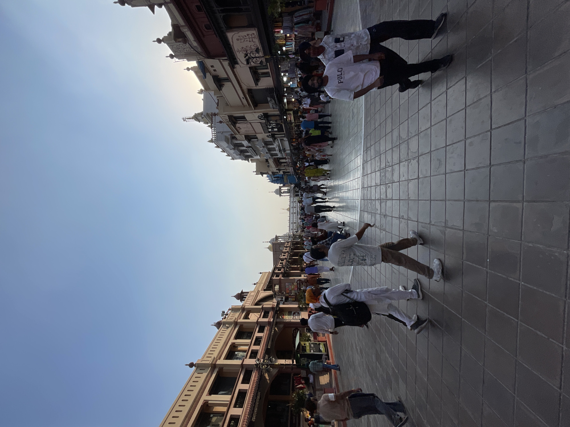 The image depicts a bustling walkway in Amritsar, India, likely leading to the Golden Temple. The scene is filled with pedestrians walking in both directions, dressed in a mix of casual and traditional Indian attire. Many individuals are wearing turbans, indicative of the Sikh community. The architecture lining the walkway features ornate buildings with arched doorways and decorative elements, suggesting historical significance. Shops selling various goods are visible along the sides of the path, with racks of clothing displayed outdoors. The ground is paved with large, square tiles, and the sky above is a clear blue with the setting sun casting a warm glow on the horizon, partially obscured by buildings. The overall atmosphere is one of active pedestrian traffic and a vibrant public space.
