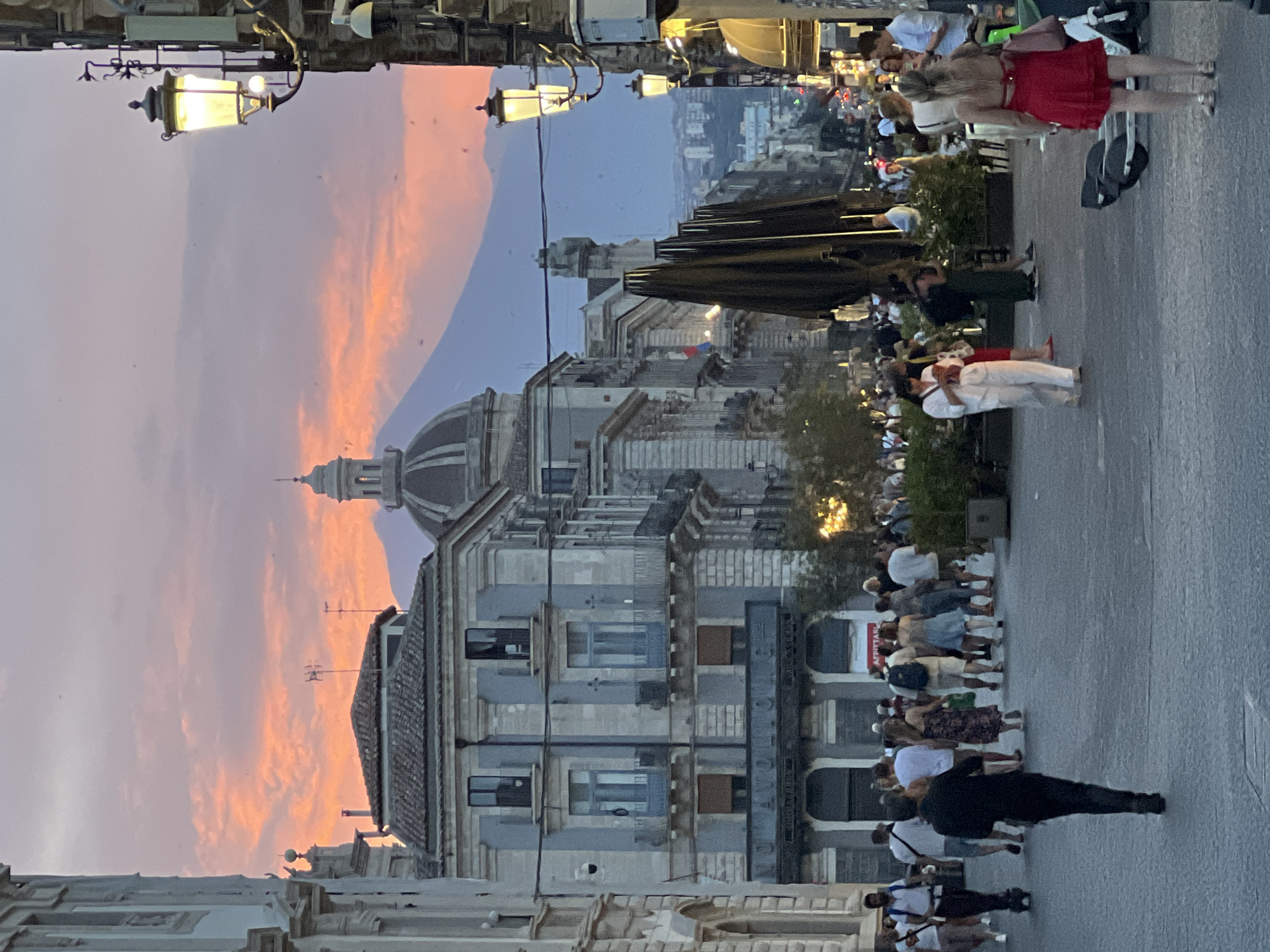 The image captures a street scene in Catania, Italy, during sunset, with Mount Etna looming in the background. The sky is painted with vibrant hues of orange and pink, indicative of twilight. In the foreground, a bustling street is filled with pedestrians walking in various directions. Some individuals are casually strolling, while others appear to be heading towards shops or restaurants lining the street.
The architecture along the street features classic European design, with ornate buildings and balconies. Notably, a building with a prominent dome is visible on the left side of the image, and streetlights cast a warm glow, illuminating the path. Several parked scooters and a bicycle suggest a mode of transportation common in the city. The presence of umbrellas and outdoor seating hints at cafes or dining establishments open for business. The overall atmosphere appears lively and vibrant, reflecting the typical evening ambiance of a European city.