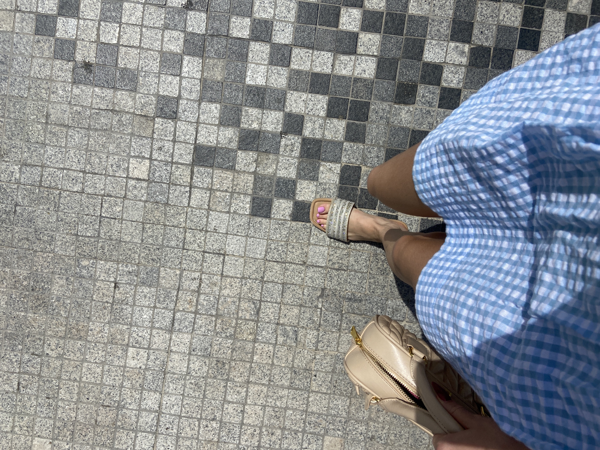 The image shows a partial view of a person standing on a pavement made of light and dark grey square stones, possibly granite, in a checkerboard pattern. The person is wearing a blue and white checkered dress and a sandal on their right foot with pink nail polish on the toes. A beige, quilted handbag is partially visible at the bottom right of the image. The location cannot be determined from the image, but the prompt suggests Dubai, United Arab Emirates. No specific actions or interactions are evident in the image.