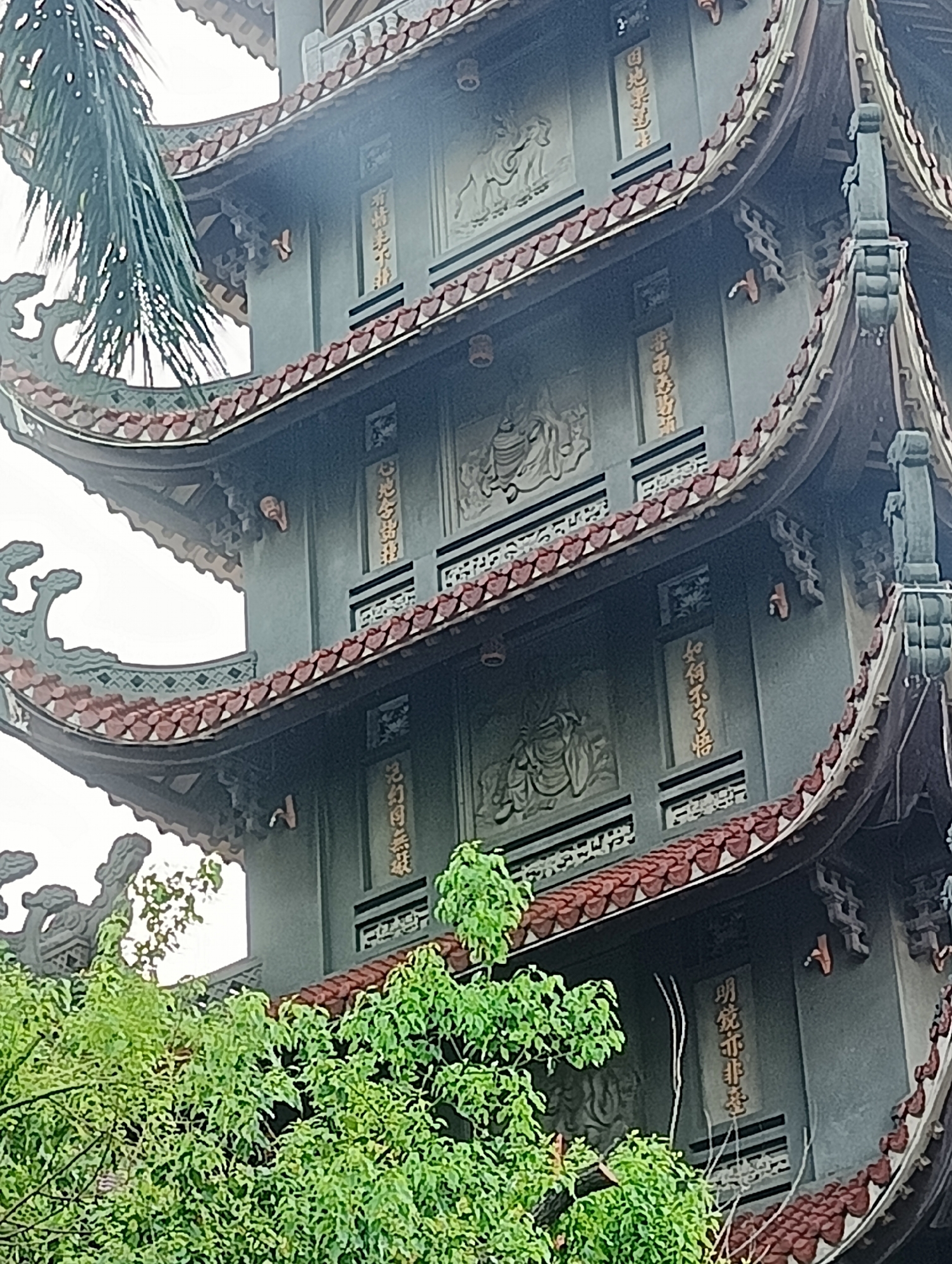 The image captures the upper sections of a multi-tiered pagoda, likely located in Thủ Đức City, Vietnam. The pagoda's architecture features curved, tiled eaves with dark red tiles and decorative green trim. The gray walls are adorned with bas-relief sculptures depicting figures, possibly deities or historical figures, and vertical inscriptions in what appears to be gold-colored script. The foliage of lush green trees partially obscures the lower portions of the pagoda. The sky is overcast and white.