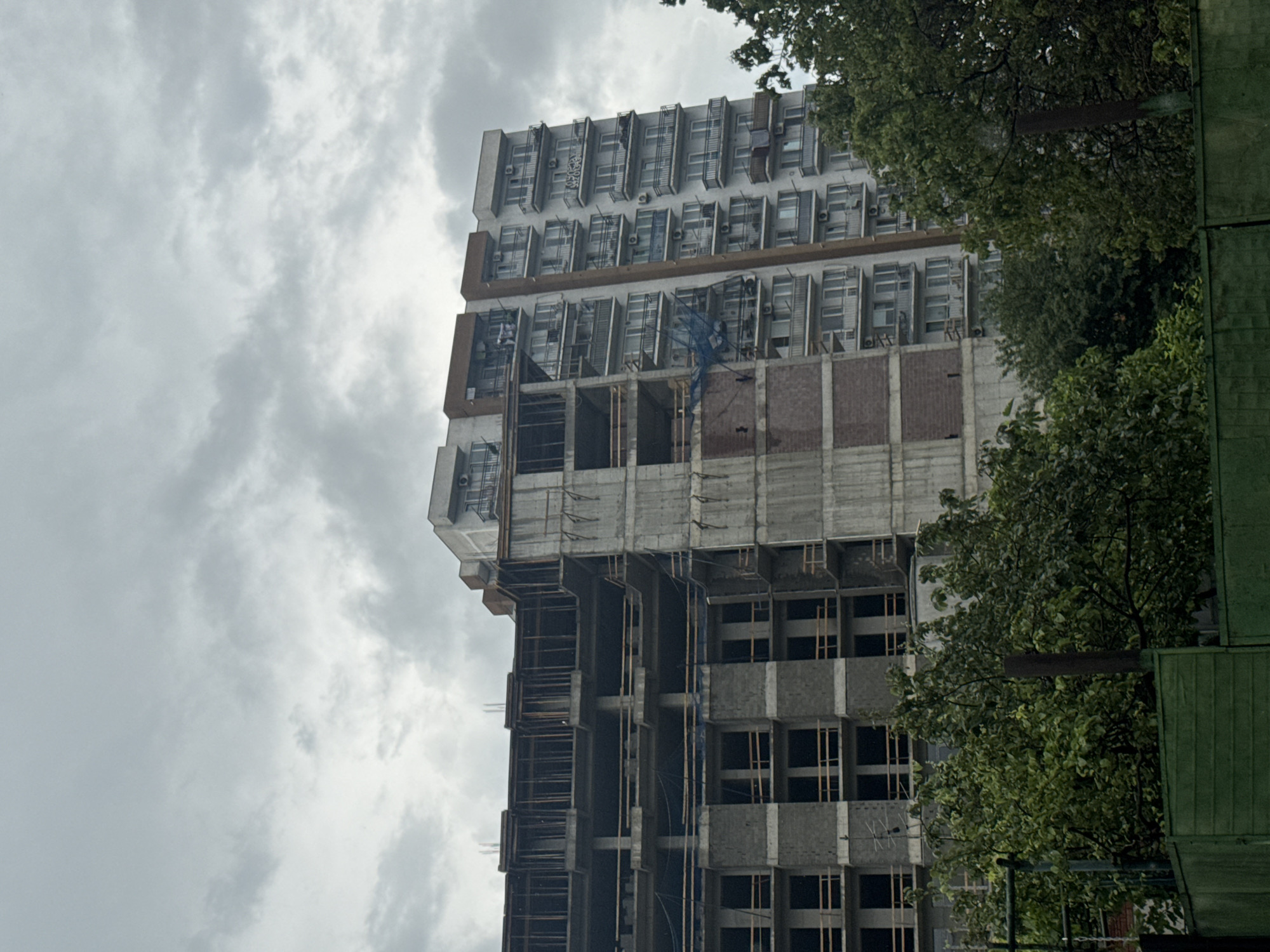The media file shows a tall, multi-story building under construction in Tbilisi, Georgia. The building has a modern architectural design, with numerous windows and balconies. Parts of the building are still in a raw concrete state, indicating ongoing construction work. Higher floors appear to be more complete, with windows installed and a facade resembling white panels. Lush green trees partially obscure the building, suggesting an urban environment with natural elements. The sky is overcast with heavy clouds, indicating a cloudy day, possibly with impending rain. There is no visible text in the media file. The overall impression is one of urban development and progress, set against a slightly somber, cloudy sky.