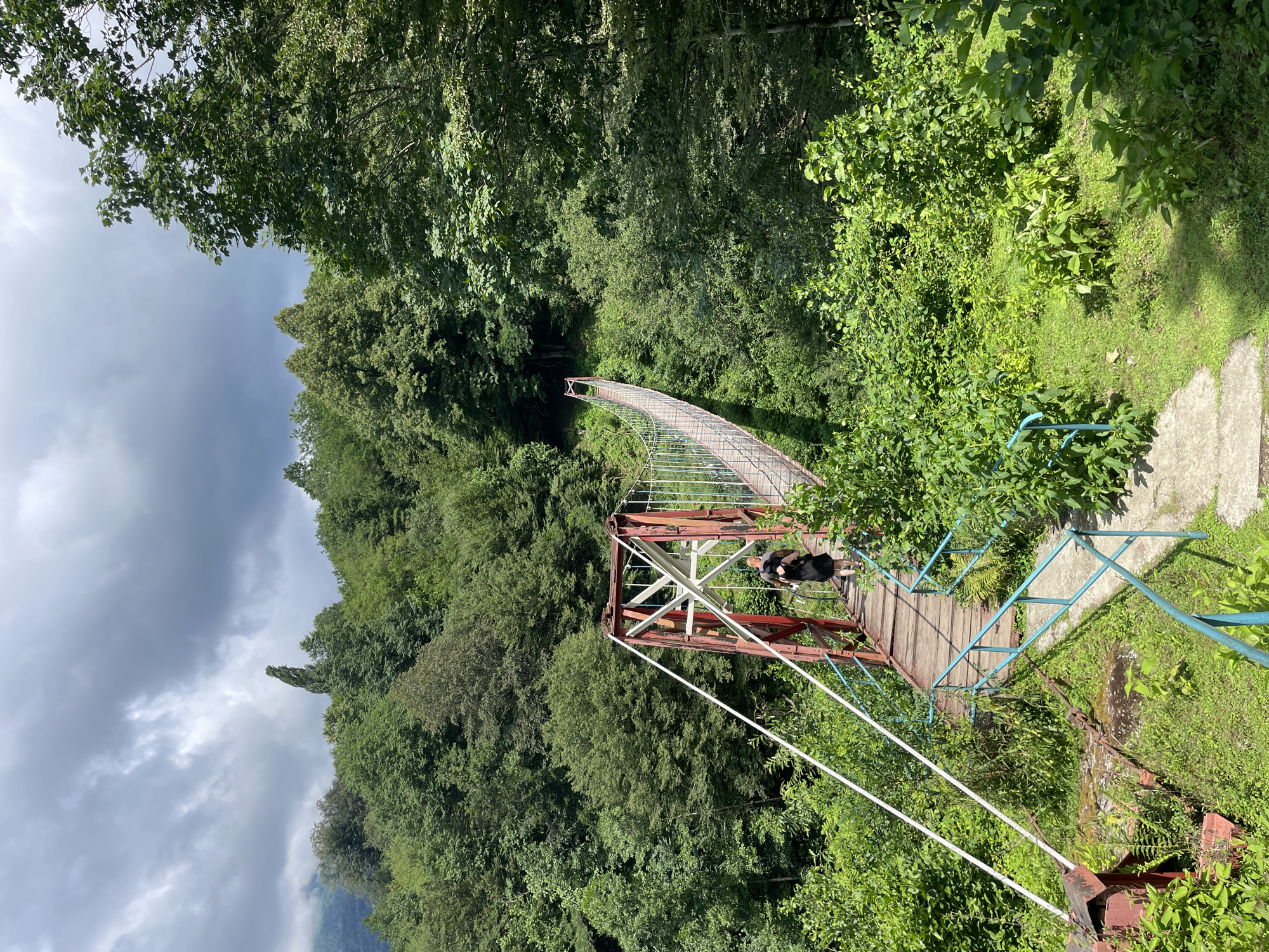 The image shows a pedestrian suspension bridge in Tqwarchal, Abkhazia. The bridge has wooden planks and is supported by red metal structures. A person wearing a black dress is walking across the bridge. The bridge spans across a lush, green, forested area with dense trees. The sky is cloudy, with dramatic light filtering through. The foreground shows overgrown vegetation and concrete steps leading to the bridge.
