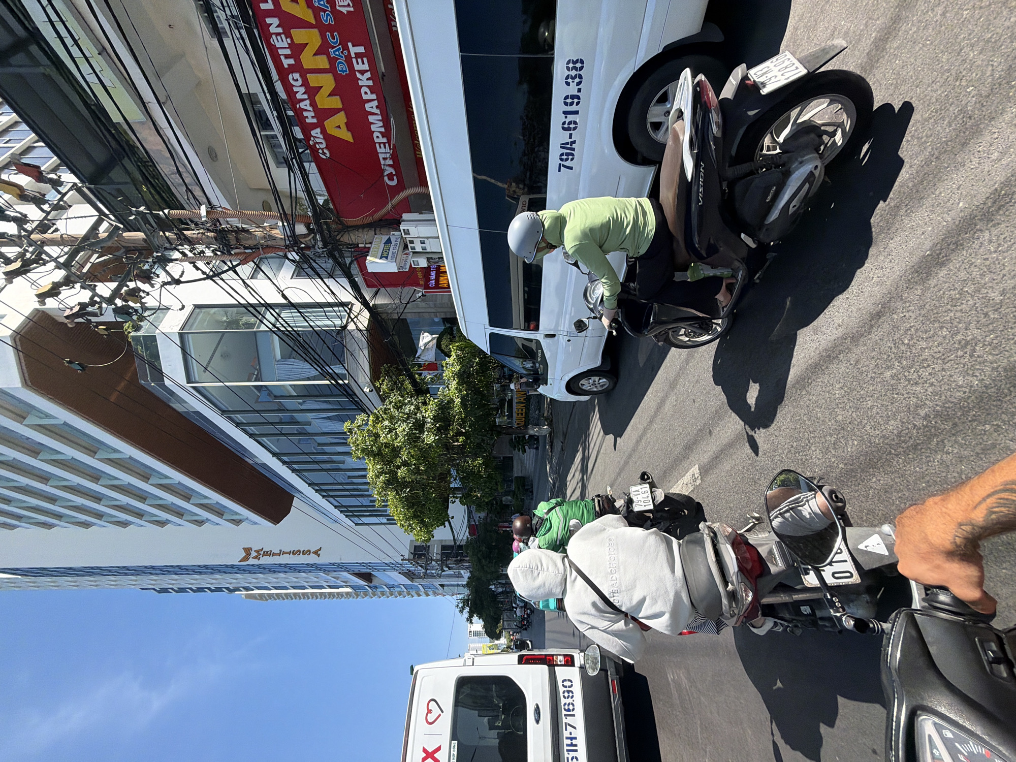 The image captures a street scene in Nha Trang Ward, Vietnam, on a bright, sunny day. The perspective is from someone riding a motorbike, looking forward. In the foreground, a hand with tattoos is visible, gripping the motorbike's handlebar.
In front of the viewer's motorbike, another motorbike is moving away, carrying two individuals. The rider is wearing a green long-sleeved shirt and a helmet, while the passenger is in a white hoodie. The passenger's hoodie has text on the back that reads "NOT BAD CHOICED." Both motorbikes are in traffic, with other motorbikes and a white van visible further ahead. The license plate of the motorbike in front is partially visible, showing "79-VA 104.61".
To the right of the road, a white van with tinted windows is driving, displaying the license plate "79A-619.38". A red sign with yellow text is visible above the van, advertising "ANNA SUPERMARKET". To the left of the road, a tall, modern building with a light-colored facade and numerous balconies dominates the skyline. The building has a prominent sign that reads "MELISSA".
The environment appears to be a busy urban street with buildings, power lines, and trees lining the sides. The road surface is asphalt. The overall impression is one of active daily life in a Vietnamese city.