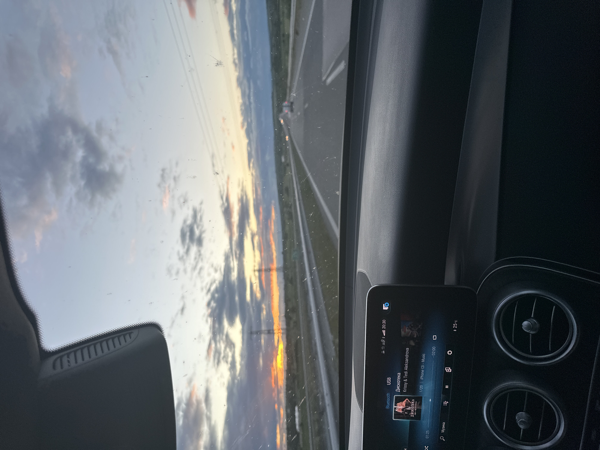 The image shows the view from inside a car driving down a highway, likely near Krum, Bulgaria, at sunset. The sky is visible through the windshield, with clouds tinged orange and pink by the setting sun. The road stretches ahead with visible lane markings, and some distant vehicles can be discerned. Inside the car, the dashboard is in focus. A car infotainment system screen shows that music is playing from a USB device, the song is titled "Дискотека" by Krissy & Tedi Aleksandrova. The temperature displayed is 25°C. Two circular air vents are visible on the dashboard to the right of the screen.