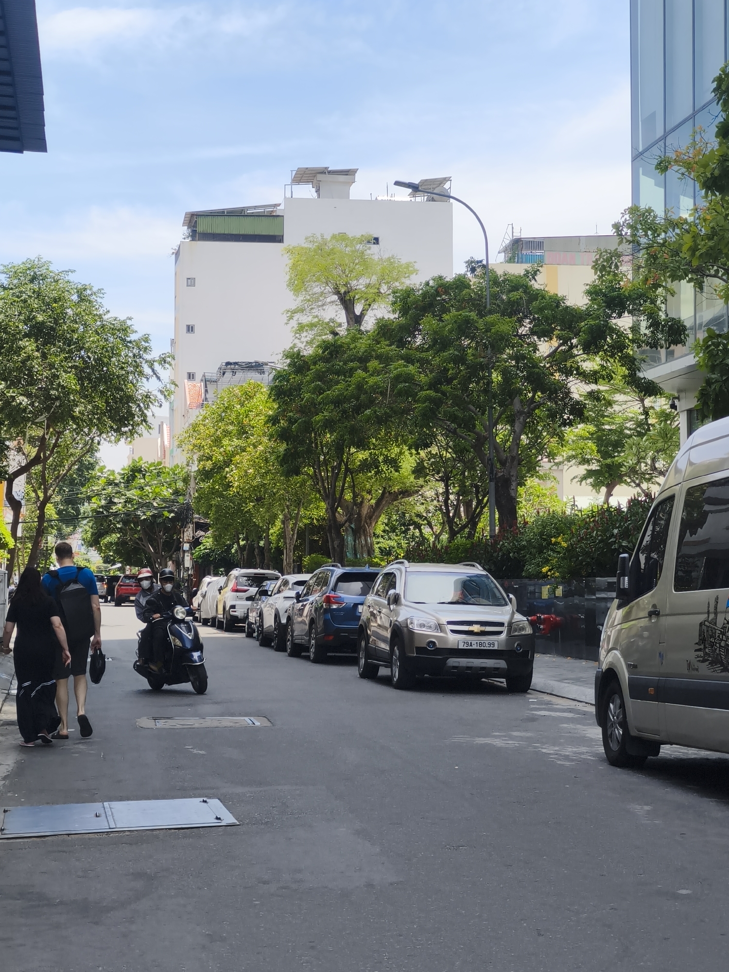 The image depicts a street scene in Bac Nha Trang Ward, Vietnam, under a partially cloudy sky. In the foreground, a paved street shows some wear and tear. On the left side of the street, two individuals, a woman in a black dress and a man in a blue shirt and backpack, are walking away from the camera. Further up the street on the left, a dark-colored scooter with two passengers wearing helmets is moving away from the camera. A line of parked cars, including SUVs and sedans, stretches along the right side of the street, most of them light-colored. The cars are parked closely together. A light-colored van is visible on the far right, partially obscuring the view of some of the parked cars. Tall green trees line both sides of the street, providing shade. Behind the trees, buildings of varying heights are visible, including a tall white building with solar panels on its roof and a modern glass-fronted building on the right. The overall impression is of a typical urban street in Vietnam with traffic and pedestrian activity.
