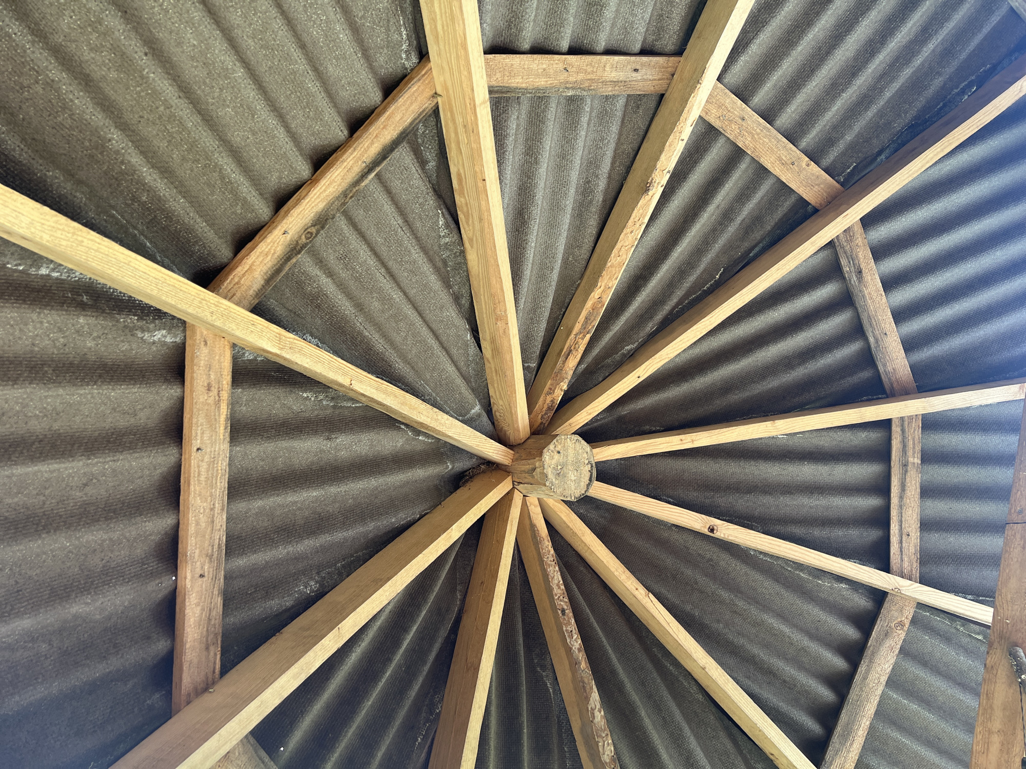 The image shows the underside of a rustic, octagonal roof structure, likely part of an outdoor shelter or outbuilding. The roof is constructed with corrugated, dark gray roofing material, possibly asbestos-cement sheets, attached to a wooden framework. The framework consists of radial wooden beams that converge at a central point where a cylindrical wooden element is visible. Additional horizontal wooden bracing pieces are also present, connecting some of the radial beams. The wood appears unfinished and weathered, with visible grain and knots. The lighting suggests an outdoor environment, with light filtering through the structure. The specific location in Norviliškės, Lithuania, is inferred from the provided prompt, but no visual cues within the image directly support this identification. There are no people or specific actions depicted; the image focuses solely on the structural elements of the roof.