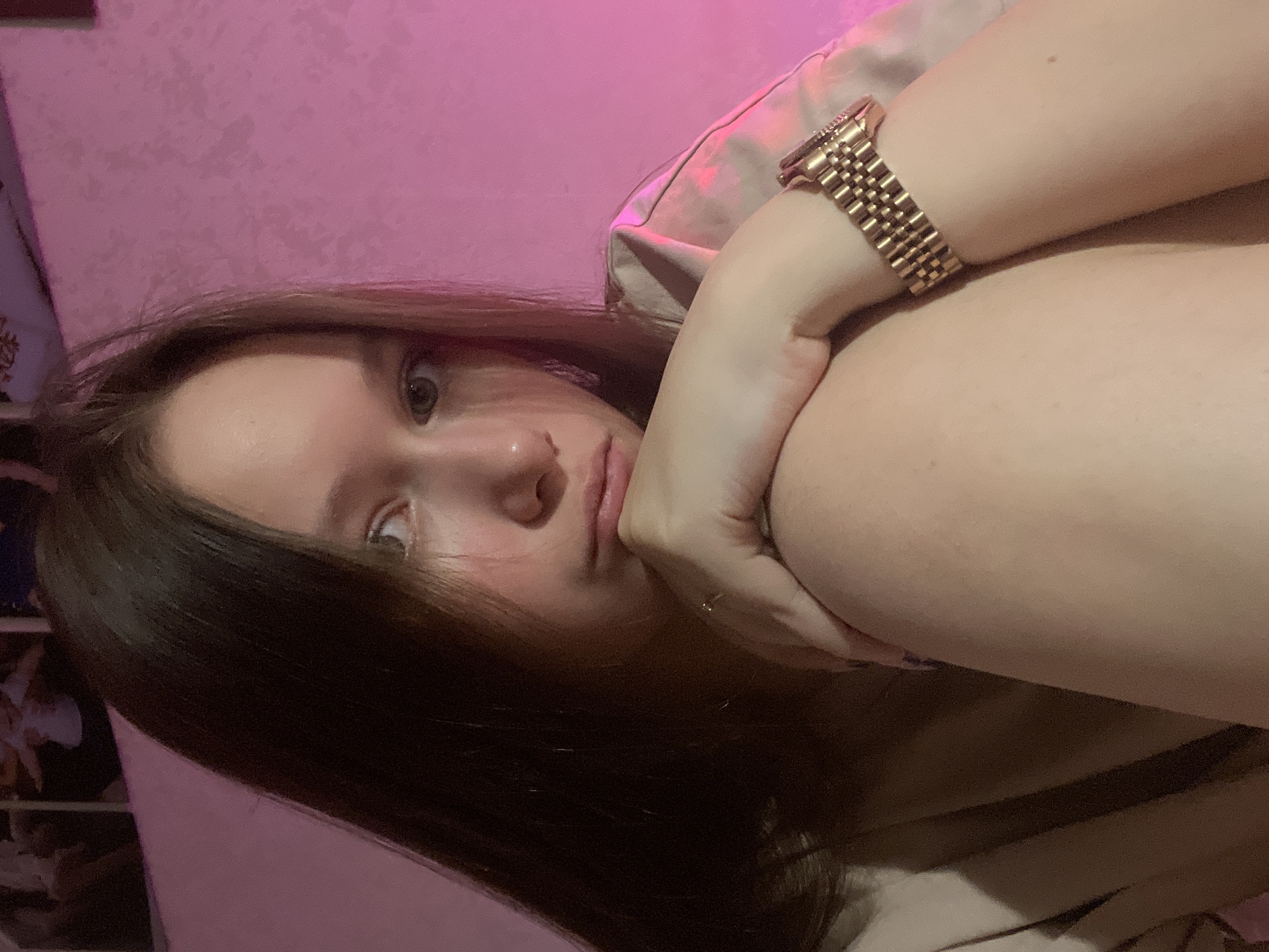 The image is a selfie of a young woman with long brown hair, leaning against a pillow. She's wearing a gold wristwatch and has her arm bent, with her hand near her face. The background is a pink wall. The photograph appears to be taken indoors. There is no indication of the location in the city of Chaikovsky, Russia.