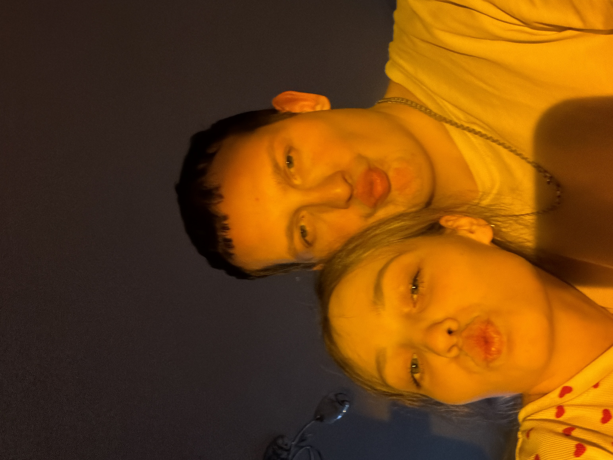 Two individuals, one male and one female, are close to the camera, their faces positioned intimately together. Both are making a puckering facial expression as if preparing to kiss. The male has dark hair, and the female has light hair. The male appears to be wearing a yellow t-shirt and a chain necklace. The female appears to be wearing a light-colored shirt with red heart patterns. The background is dimly lit and nondescript. There is no information given in the image to verify the location is Chaikovsky, Russia.