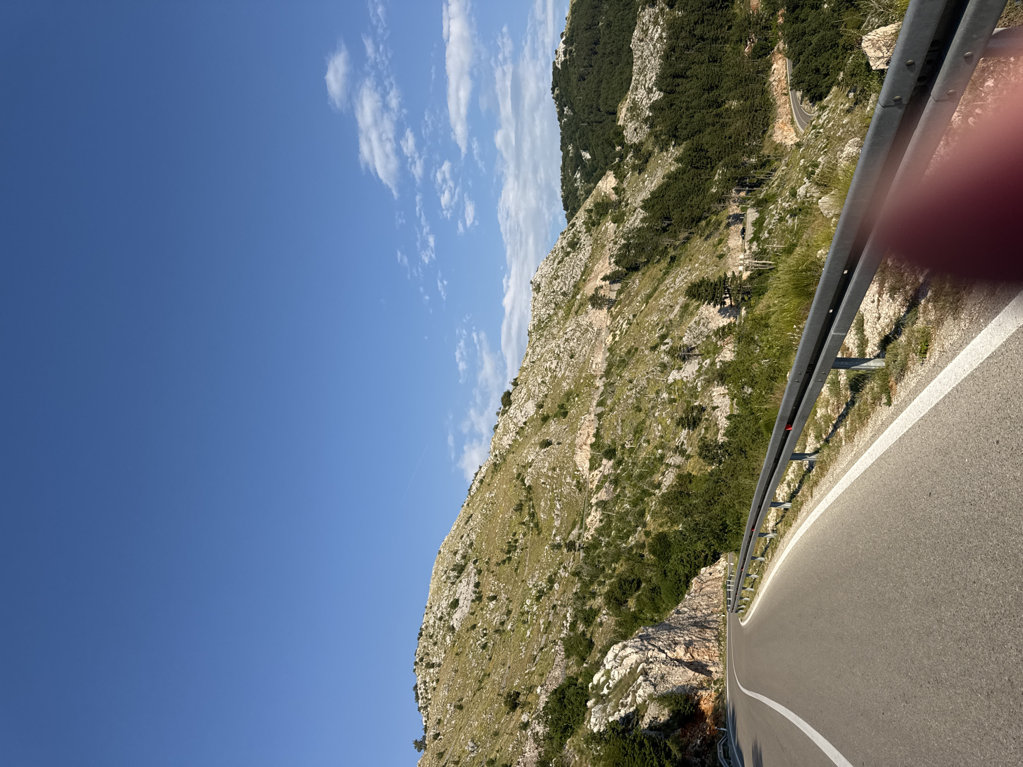 An asphalt road with white lane markings curves through a mountainous landscape under a clear blue sky with scattered white clouds. A metal guardrail lines the right side of the road. The mountainside to the right is covered in sparse green vegetation, rocks, and trees, with a winding road visible further up the slope. The mountainside to the left features rocky cliffs and more vegetation. The scene appears to be in a rural, mountainous area. The image was taken in Bukovica, Montenegro.