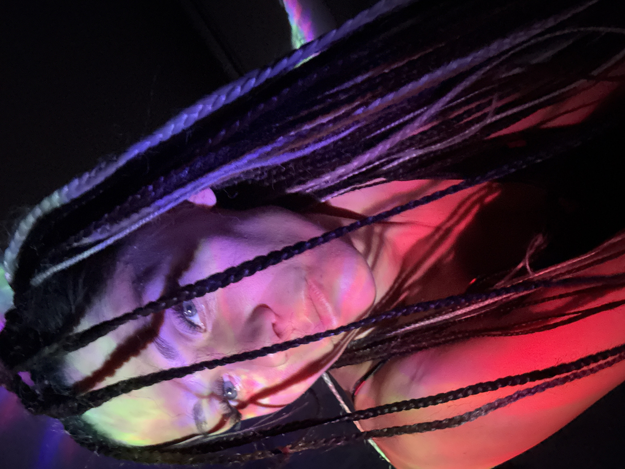 A close-up portrait features a person, likely female, with long, dark braided hair, partially obscuring her face. Her head is tilted, and she gazes directly at the viewer with clear, light-colored eyes. The scene is characterized by dramatic, multi-colored artificial lighting, casting vibrant hues across her skin and hair. Shades of purple and pink/red are particularly prominent on her face and neck, with touches of green and yellow light also visible, creating a dynamic and atmospheric effect.
The background is dark, emphasizing the subject and the interplay of light and shadow. The braided hair, possibly dreadlocks or box braids with some lighter sections, drapes across her face, forming striking lines and adding depth to the composition. Her expression appears neutral, direct, and perhaps contemplative. The colored illumination suggests an indoor setting such as a club, concert, or a venue with artistic lighting, implying the event takes place at night or in a controlled lighting environment. The image is angled, contributing to its artistic and immersive feel. No specific activity beyond posing for the camera is discernible, and no text is visible within the frame. The portrait was captured in Rusanovo, Russia.