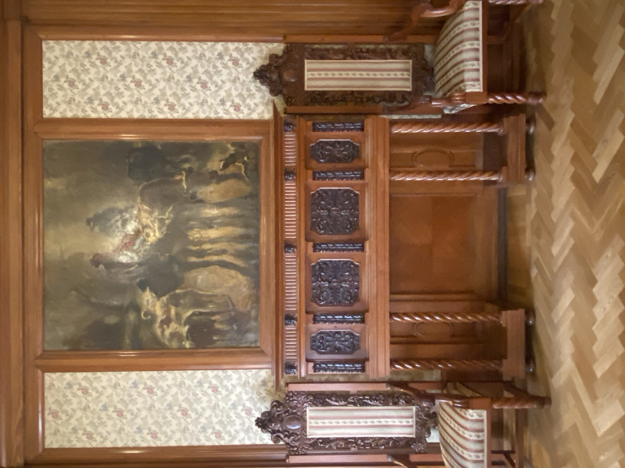 The image depicts a richly decorated room, likely a drawing-room or reception area, featuring ornate wooden furniture and a large painting. Two elaborately carved dark wood chairs with upholstered seats and backrests, featuring a striped pattern in shades of cream, red, and pink, flank a substantial wooden fireplace surround. The fireplace surround is made of polished wood and displays intricate carvings, including decorative panels and fluted columns. Above the fireplace, a large framed painting is hung, depicting a scene with figures on horseback and animals in what appears to be a woodland setting. The walls are adorned with patterned wallpaper featuring a delicate floral design in muted colors. The floor is covered in a herringbone pattern of polished wooden planks. The overall impression is one of opulence and historical grandeur. The scene is likely located in a palace or historic house museum in Saint Petersburg, Russia, given the architectural style and furnishings.