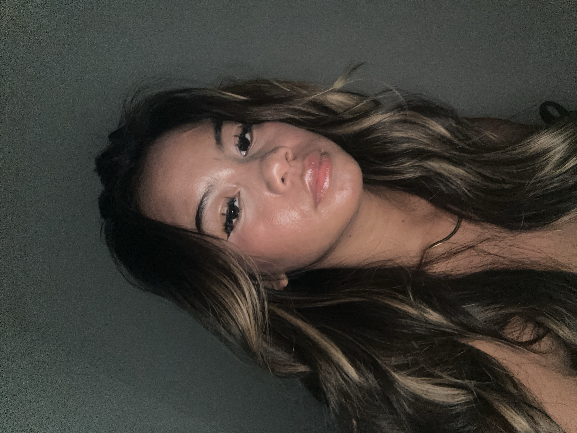 A young woman with long, wavy brown hair with blonde highlights is shown in a close-up, low-angle selfie. She has light to medium skin tone, dark eyes, and glossy lips. The background is a dark, solid color, possibly a wall. The environment provides no visual information to identify the location as Sukawati, Indonesia.
