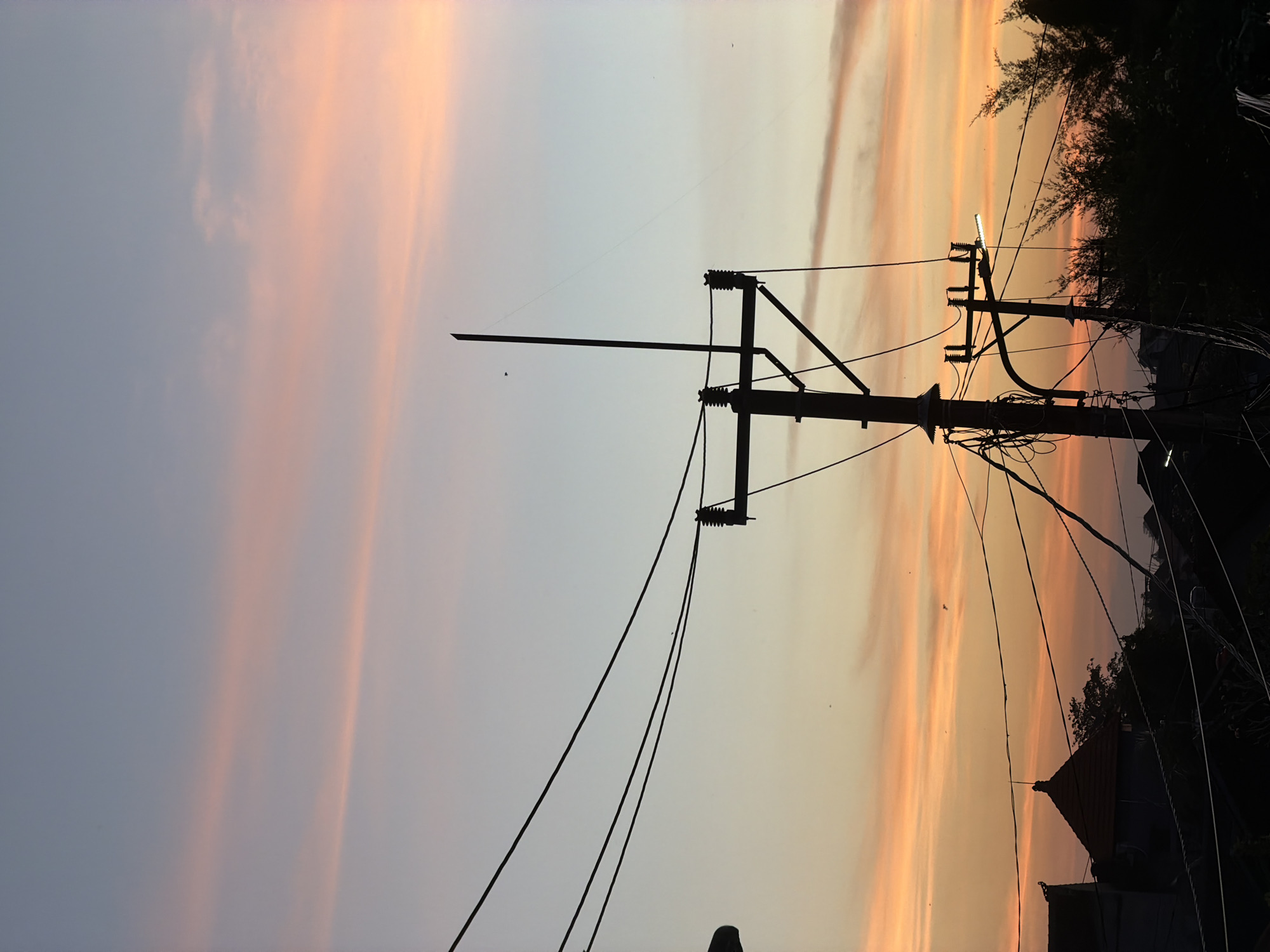 The image captures a twilight scene in North Kuta, Indonesia. A silhouette of a utility pole dominates the foreground, with electrical lines extending towards the horizon. The sky displays a gradient of colors, transitioning from light blue to shades of orange and pink, suggesting either sunrise or sunset. Silhouettes of trees and a building are visible against the brighter sky on the right side of the image.