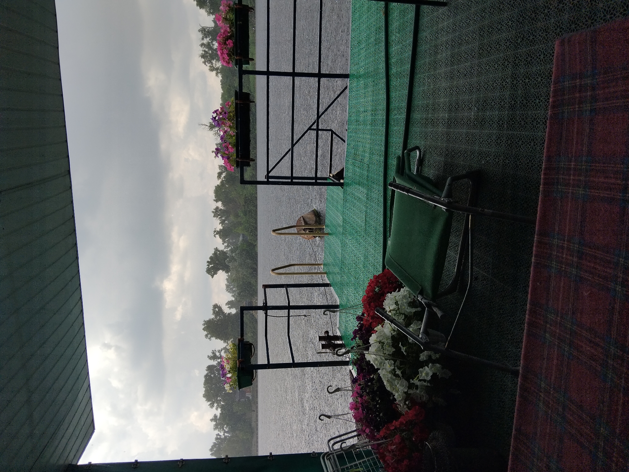 The image depicts a rainy day on a boat or a pier overlooking a body of water, likely the Dnipro River, in Kyiv, Ukraine. The foreground shows a covered seating area with a plaid blanket draped over a surface. A green chair is placed on a textured green mat. Hanging baskets filled with colorful flowers, including red, white, and purple petunias, are visible. In the midground, there is a railing and a green walkway leading to a dock or platform. Two planter boxes with pink and purple flowers are mounted on the railing. The background reveals a treeline and a cloudy, overcast sky, with visible raindrops hitting the water. No people are present in the scene.