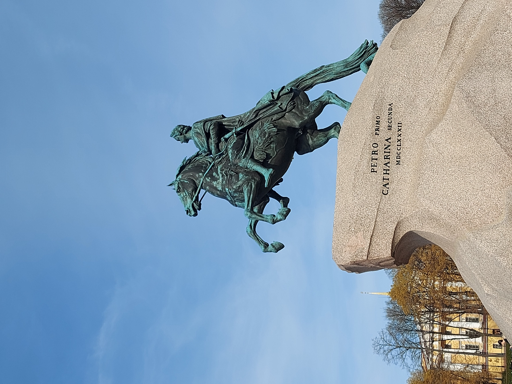 The image shows the Bronze Horseman, a statue of Peter the Great in Senate Square in Saint Petersburg, Russia. The statue depicts Peter the Great on horseback, rearing up, on top of a massive stone pedestal. The inscription on the stone reads "PETRO PRIMO CATHARINA SECUNDA MDCCLXXXII" (To Peter the First from Catherine the Second 1782). The background shows a clear blue sky and buildings with an architectural style typical of Saint Petersburg. The statue and its setting suggest a historical and cultural landmark.
