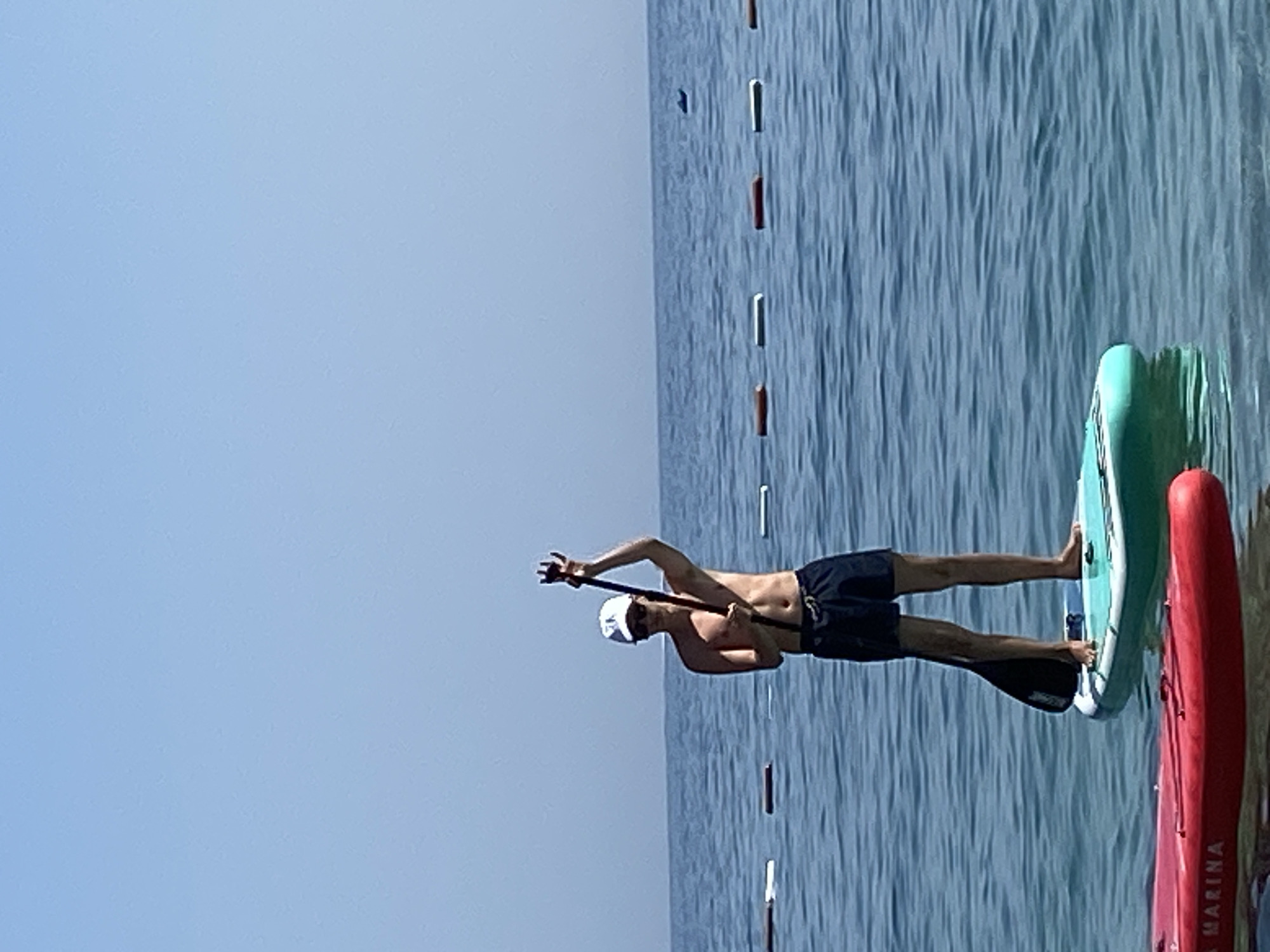 A shirtless man wearing a white cap and dark swim shorts stands on a turquoise paddleboard in the calm, blue sea. He holds a paddle, its blade submerged in the water, in his right hand. The man is looking forward and appears to be in motion. In the foreground, a red kayak is partially visible. In the background, a line of buoys stretches across the water, separating a swimming area from the open sea. The sky is clear and blue. The scene is set in the city of Prijevor, Montenegro.