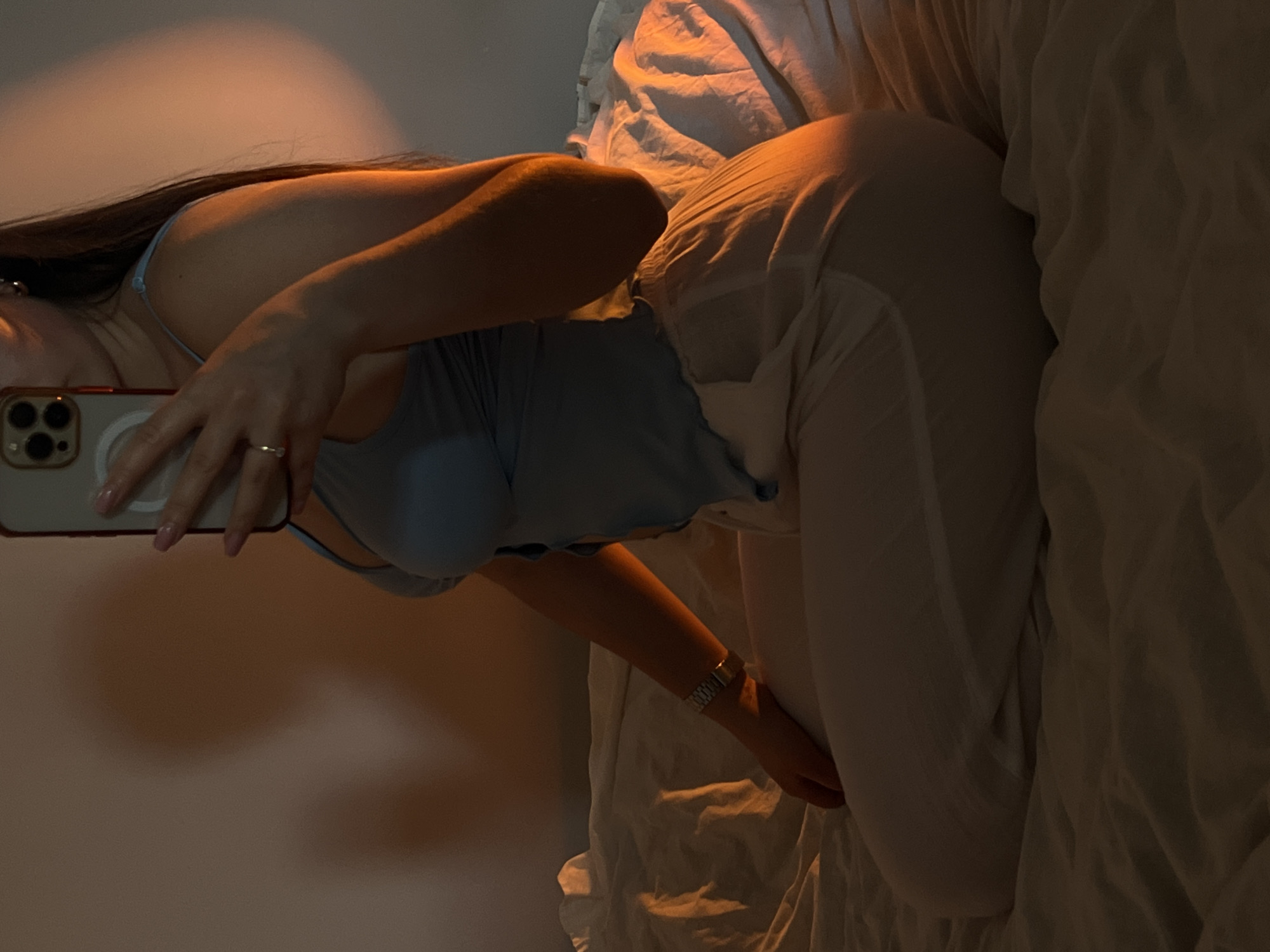 A woman takes a photo of herself while lying on a bed. She is wearing a light blue top and white pants. Her hair is dark brown. She is holding a smartphone with her left hand to take the photo. The background is a wall with soft lighting. The bed has white sheets and pillows. The location is unknown and could not be determined to be Ternopil, Ukraine.