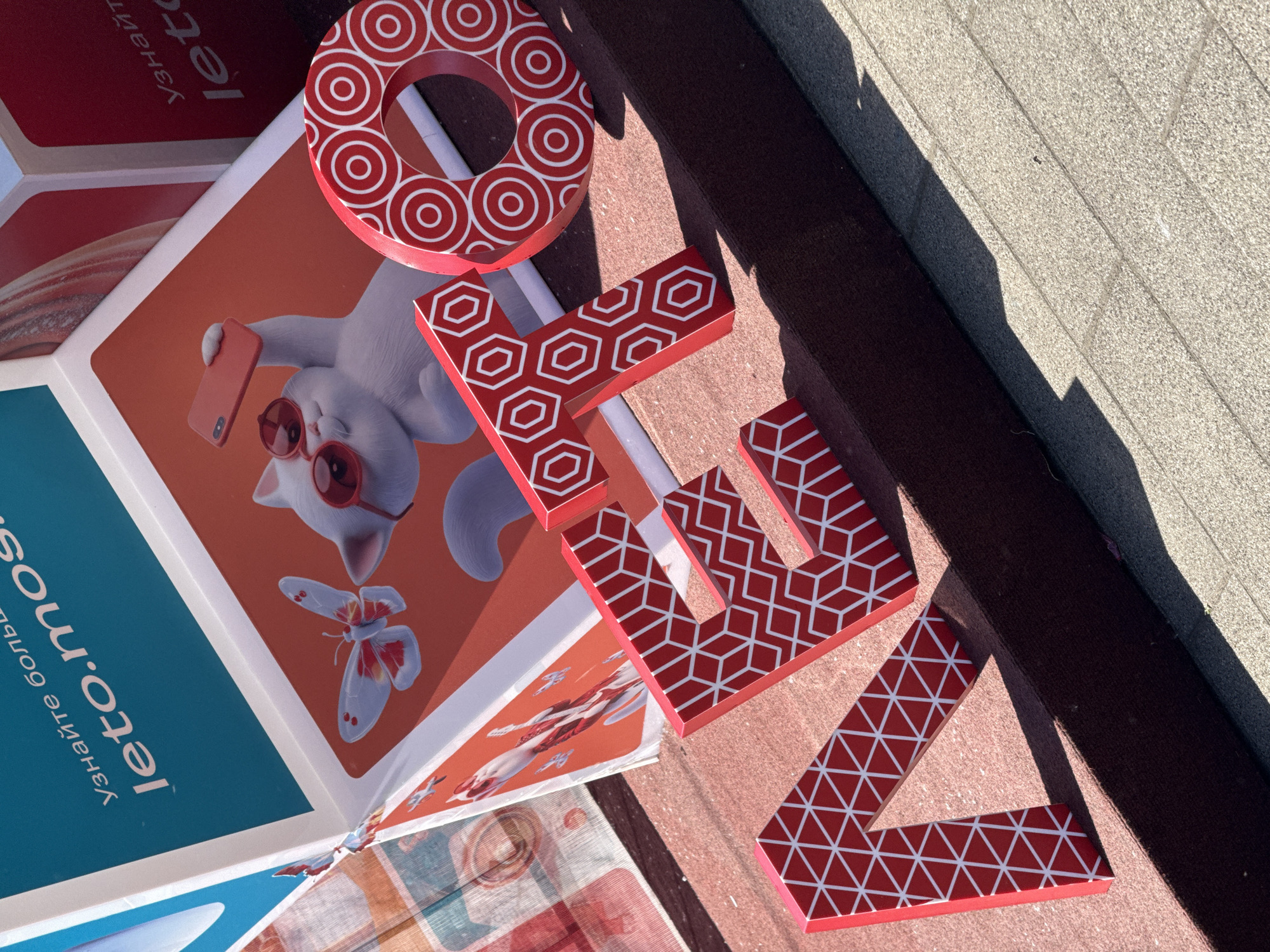 This image depicts an outdoor promotional installation for the "Leto v Moskve" (Summer in Moscow) program, located in a public space within Moscow, Russia. In the foreground, large, three-dimensional red letters spell "ЛЕТО" (LETO). Each letter features a distinct white geometric pattern: "Л" with triangles, "Е" and "Т" with hexagons, and "О" with concentric circles. These letters rest on a dark red, textured base, casting shadows onto the base and the light grey, horizontally-sectioned paved ground in the lower right.
Behind the letters, several angled display panels are visible. A prominent blue panel in the upper left corner features white text: "Узнайте больше" (Learn more) and the URL "leto.mos". Adjacent to and below this, an orange-red panel showcases a white cartoon cat wearing red sunglasses and holding a red smartphone in a selfie pose. A white and red butterfly is depicted near the cat. Partially visible further back and to the left are additional panels with graphics, including stylized red and white koi fish. The scene is brightly lit by direct sunlight, indicating an outdoor, daytime setting. There are no people visible in the frame.