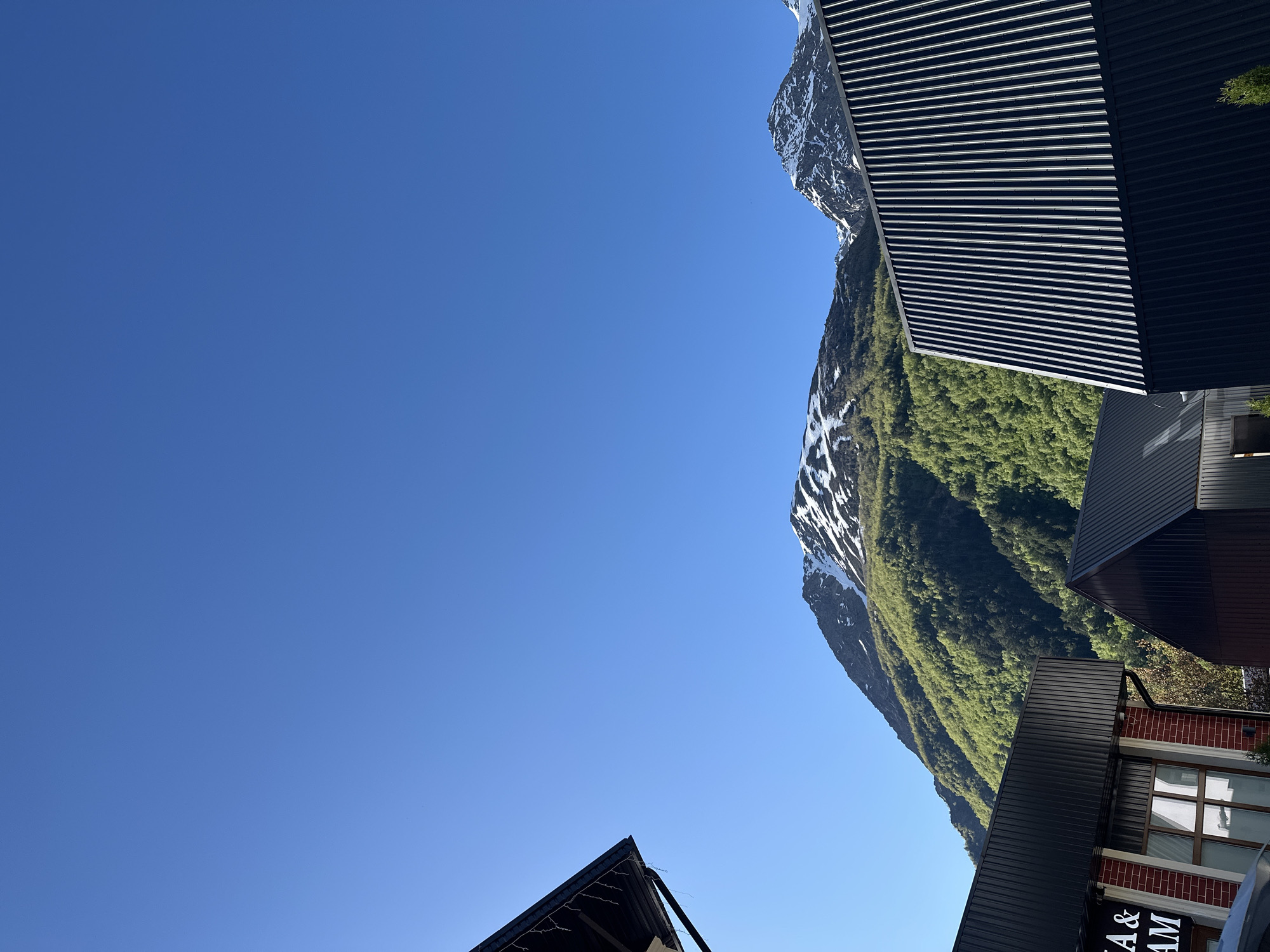 The image depicts a mountainous landscape under a clear blue sky. In the foreground, there are several modern buildings with dark, corrugated metal roofs. One building on the left has a brick facade and a sign with white lettering that reads "A & AM". A green slope covered in trees rises behind the buildings, with patches of snow visible on the higher mountain peaks in the background. The scene appears to be a view of Terskol, Russia, a mountainous region known for its natural beauty. No people or animals are visible in the image.