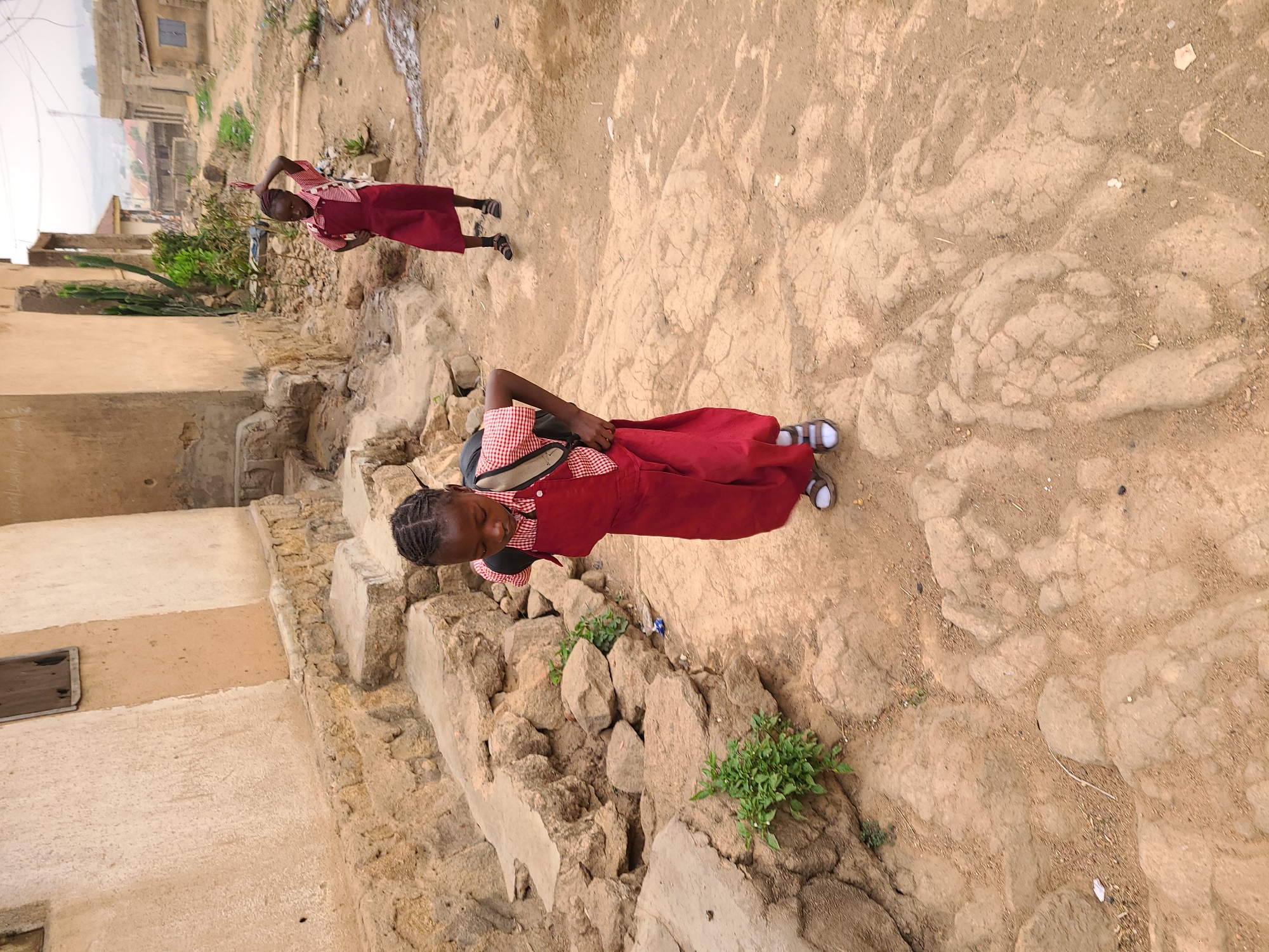 Two girls in red school uniforms and sandals are walking on a cracked, dirt path in Jos, Nigeria. One girl is closer to the camera and wearing a backpack. The background includes buildings with tan walls and roofs, and greenery growing among the structures and along the path.