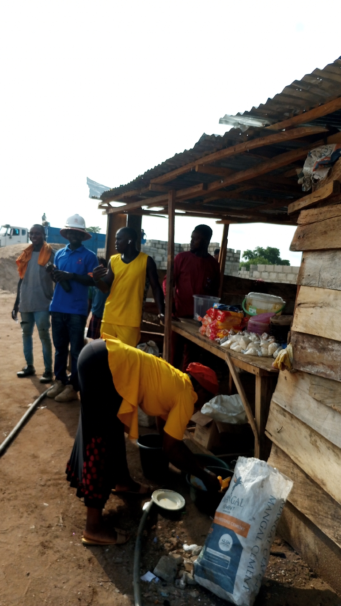 The image shows a group of people gathered around a makeshift stall, likely a small business, located outdoors in an urban environment. A woman in a yellow shirt and black skirt is bending down near the stall, possibly washing something in a bucket. Several men are standing nearby; one is wearing a hard hat and blue shirt, while others are in casual attire. The stall has a corrugated metal roof and wooden supports, with various items displayed on the countertop, including packaged goods and containers. A bag of "MANGAL CEMENT" is on the ground near the stall. In the background, there are construction materials and a piece of heavy machinery.