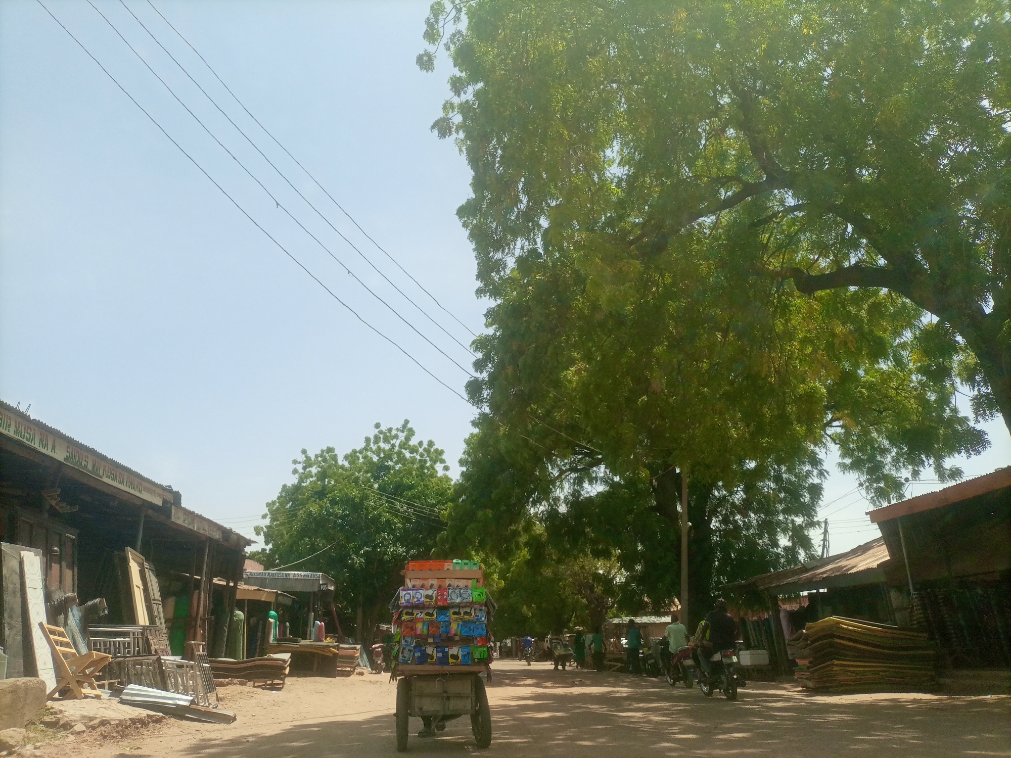 This is a street-level, eye-level shot of a dusty road in Arawa, Nigeria. On the left, a row of single-story market stalls with corrugated metal roofs line the road, displaying furniture and construction materials like wooden doors, chairs, and metal frames. On the right, more stalls are visible, with stacks of what appear to be mattresses or large cushions. A man is pushing a two-wheeled cart laden with colorful boxes, possibly of goods for sale, down the middle of the road. Several motorcycles are parked on the right side of the road, and a few people are seen near the stalls. Large, leafy trees cast shadows on the road and buildings. Overhead, electrical wires run diagonally across the pale blue sky. The overall impression is a bustling local market scene on a sunny day.