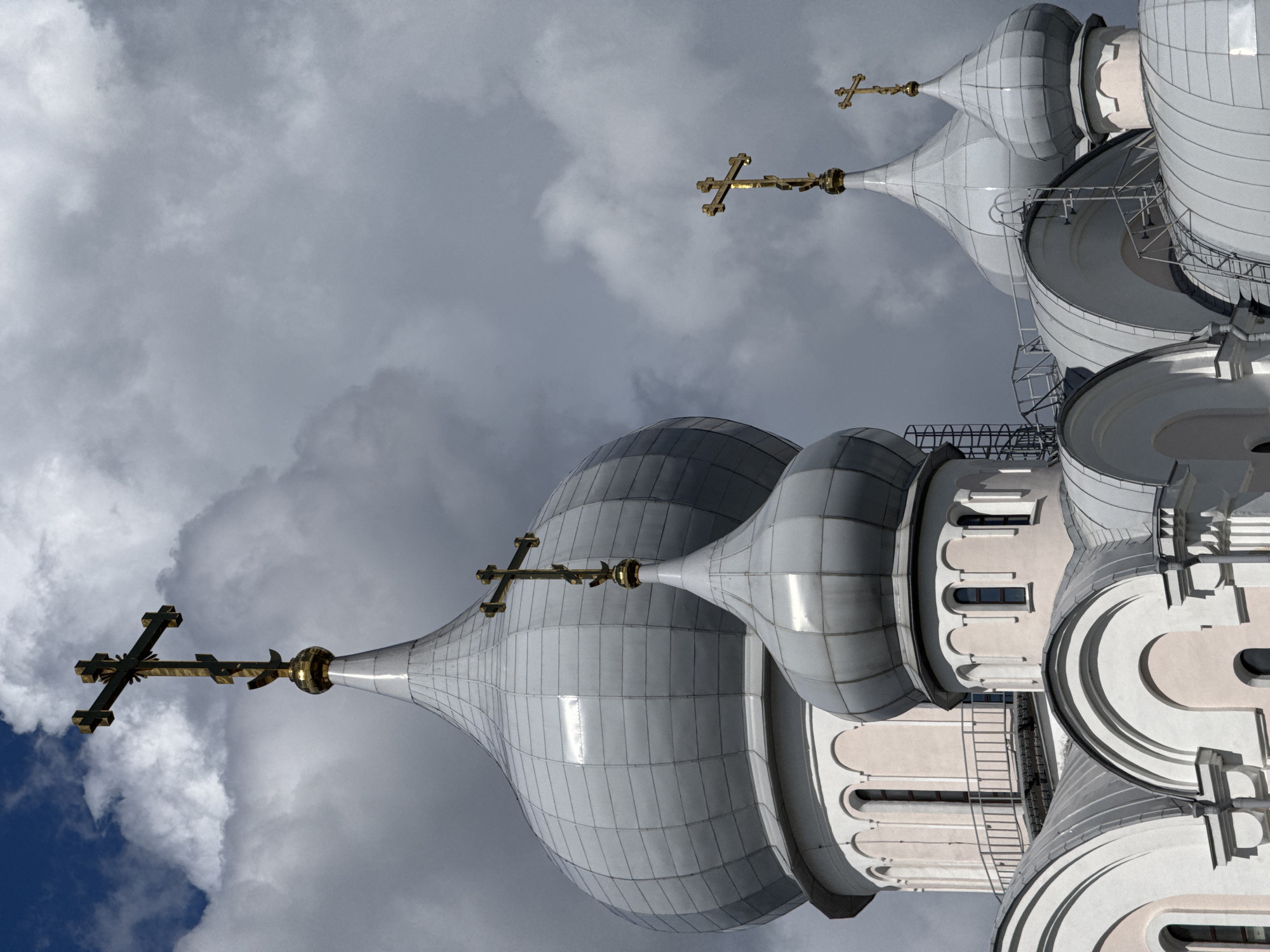 The image shows a close-up of the domes and crosses of an Orthodox Christian church, set against a dramatic, cloudy sky. The domes are large and silver, with a ribbed texture. Several golden crosses are visible atop the domes. The architectural style suggests a Russian Orthodox church. The specific building is the Church of the Resurrection of Christ on the Blood in Saint Petersburg, Russia, identified by its distinctive onion domes and ornate crosses. The lighting is dramatic, with shafts of light illuminating parts of the church and the clouds. No people are visible in the frame. The perspective is from below, looking up at the structures.