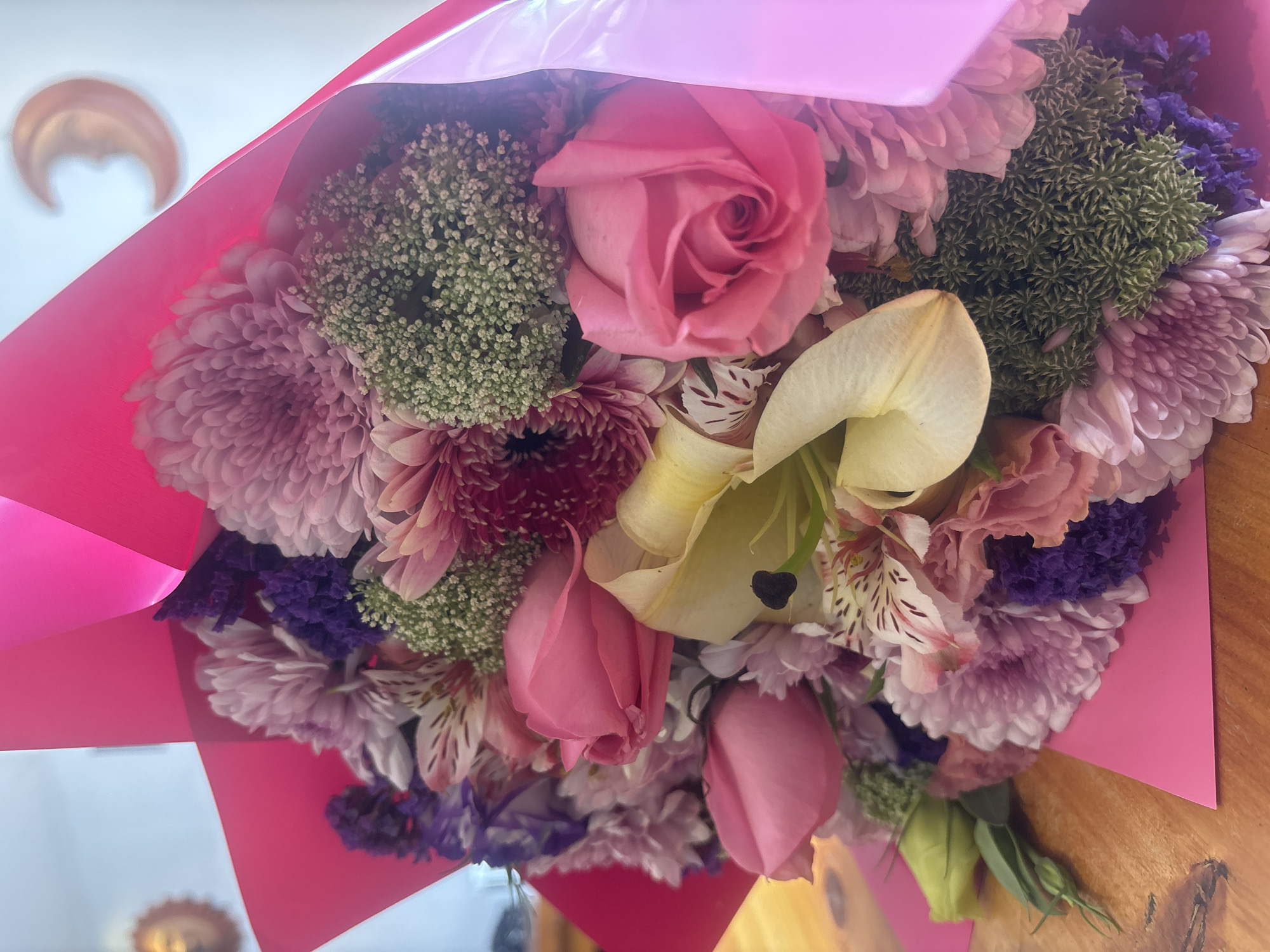 A bouquet of flowers, predominantly pink, is presented wrapped in pink paper. The arrangement includes roses, lilies, chrysanthemums, and other filler flowers with light green and dark purple hues. The flowers are arranged densely, with the lilies and roses centrally positioned. The scene is indoors, with a wooden surface visible beneath the bouquet. Parts of the setting show a glimpse of what is likely the inside of a house, with golden decorations, which suggest the location is in Puebla, Mexico, a city known for its decorative arts. No people are visible in the image.