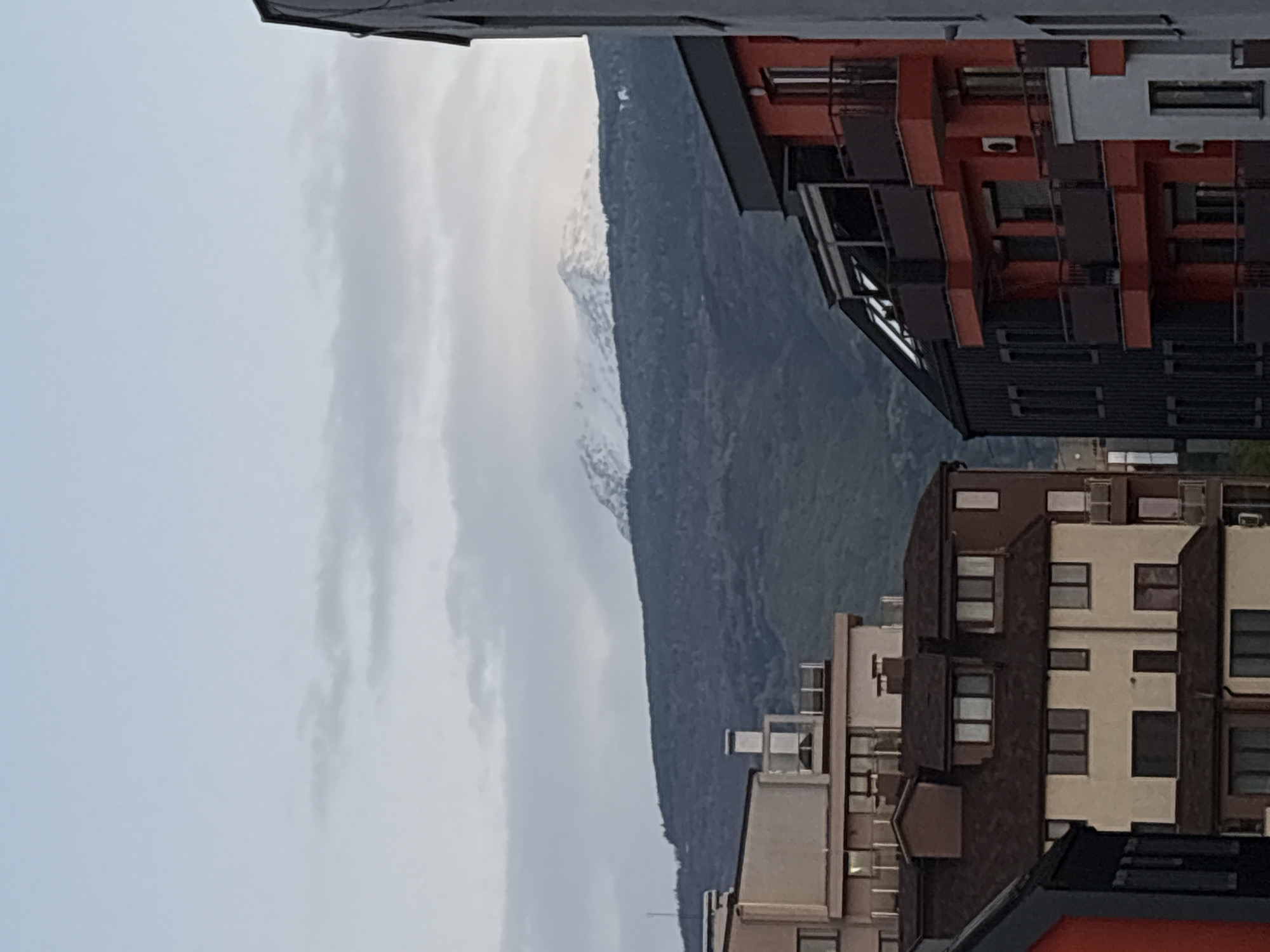 The image shows a partial view of a mountainous landscape with buildings in the foreground. The mountain is predominantly gray and green, with hints of snow near the top. Overcast sky is visible to the left of the mountain. On the right, two buildings are partially visible. The closest has a facade with a mix of orange, grey, and black elements. The other building has a brown and white facade. It is unknown if this is in Sofia, Bulgaria. There are no people visible.