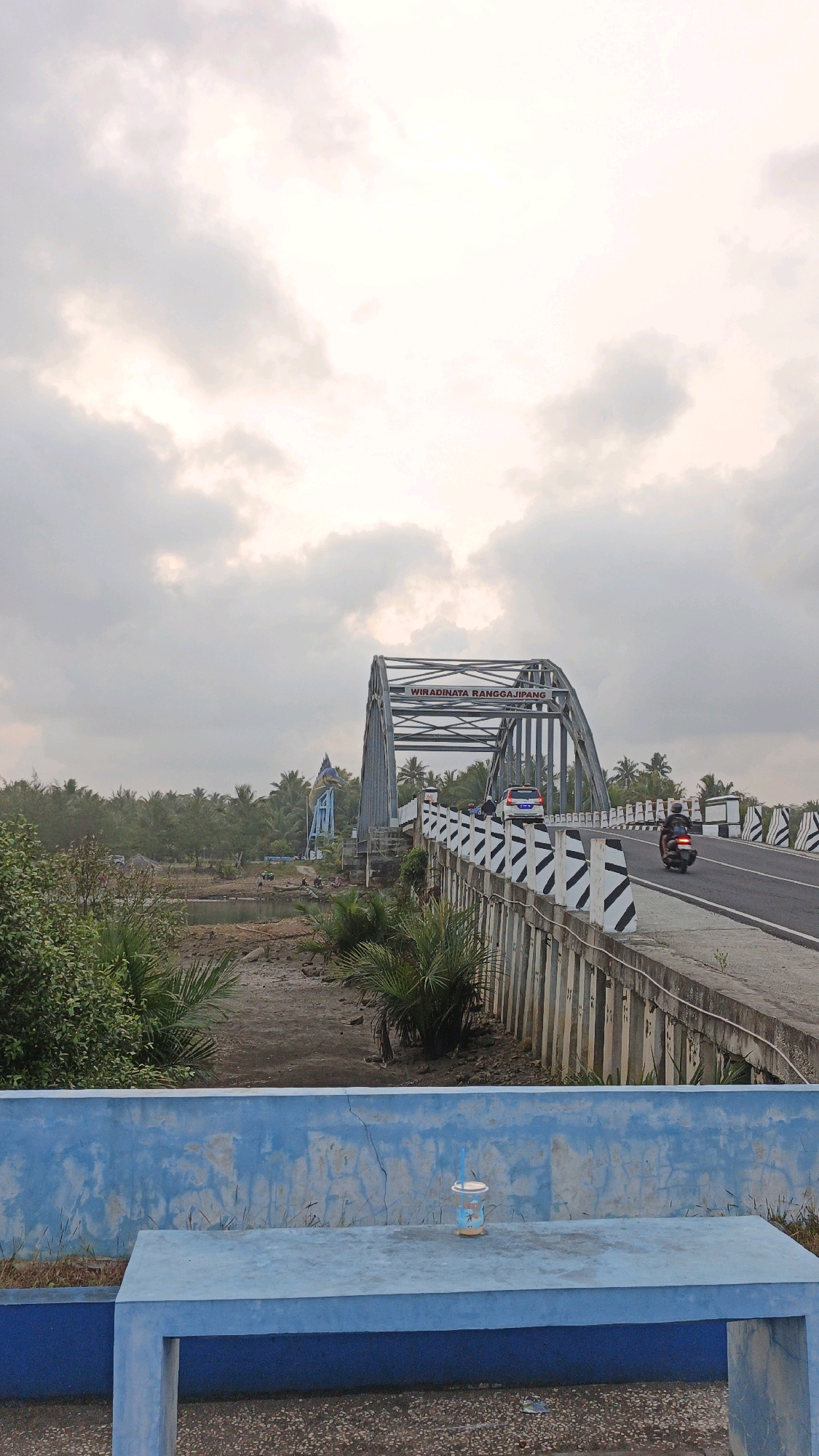 This image depicts the Wiradinata Ranggajipang Bridge in Pangandaran, Indonesia. The bridge is a steel arch structure spanning over a dry riverbed or marshy area. A road runs across the bridge, with vehicles, including a car and a motorcycle, in motion. The bridge's railing is decorated with black and white diagonal stripes. On the left side of the bridge, there is a body of water or mudflat with sparse vegetation. Further in the background, a dense forest of palm trees stretches across the horizon. The sky is overcast with clouds, suggesting either dawn or dusk. In the foreground, a bright blue concrete bench and wall are visible, with a plastic cup containing a light-colored beverage and a blue straw placed on the bench. The overall scene is one of a functional bridge in a tropical, coastal environment.
