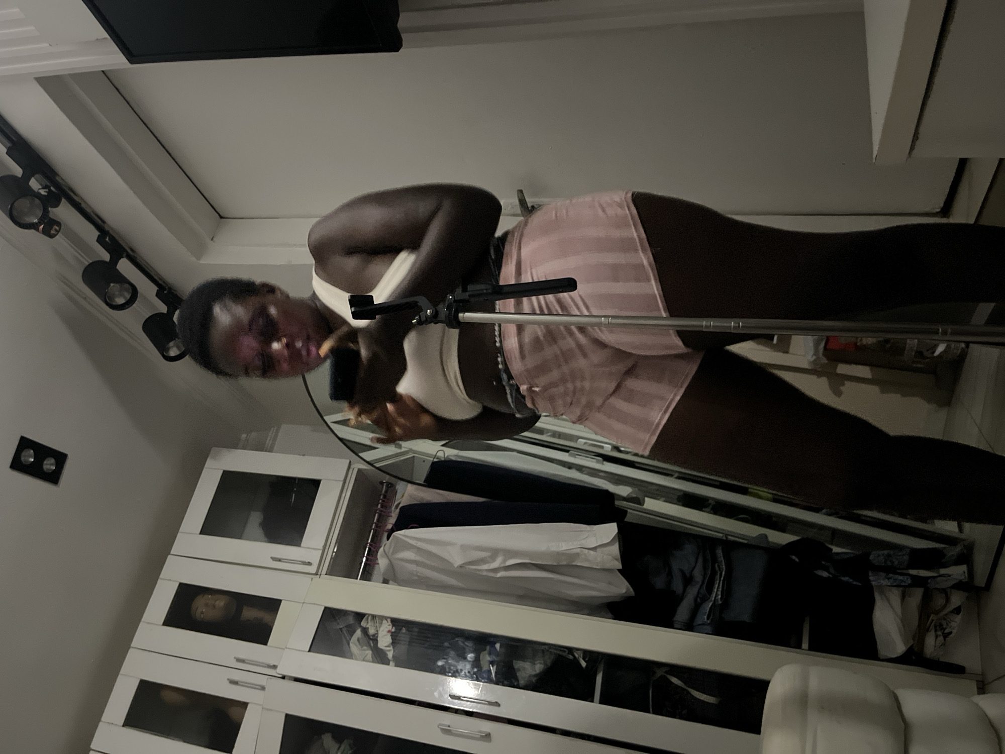 A full-body shot captures a young Black woman standing in a room, likely a bedroom or dressing area, with a large open closet to her left. She is looking down at her phone, which she holds in her right hand. A small black tripod stand is positioned in front of her, and she appears to be in the process of taking a selfie or recording a video.
The woman is wearing a white tank top and light pink striped shorts. Her hair is pulled back. The closet behind her is filled with clothing on hangers and folded items on shelves. To the right of the frame, a white door and a corner of a white dresser are visible, along with a dark-colored television mounted on the wall. Above the woman, track lighting with multiple bulbs is installed on the ceiling. The room is dimly lit, with the primary light source appearing to come from the phone and overhead lighting. The overall impression is a personal space, possibly within a residential setting in Abuja, Nigeria, given the context of the request.