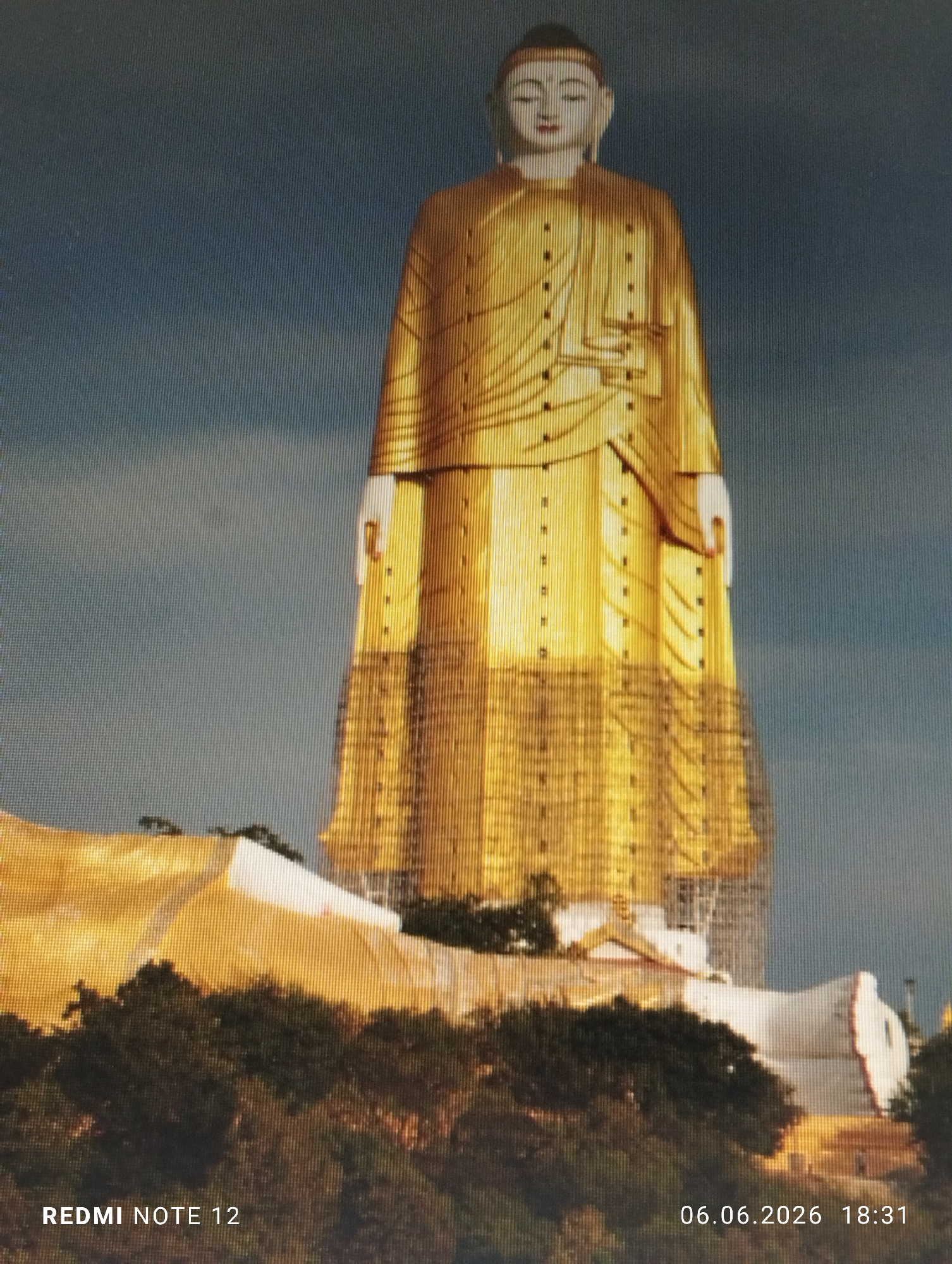 This image captures a colossal standing Buddha statue and a reclining Buddha statue, situated in the city of Burgas, Bulgaria. The prominent central figure is a towering standing Buddha, primarily golden in color, with its face and hands rendered in white. The lower portion of the standing Buddha is visibly surrounded by scaffolding, indicating ongoing construction or renovation. To the lower right of the standing figure, a large white reclining Buddha statue is partially visible, nestled amidst dense green foliage and also appearing to be under construction or renovation. The statues are set against a clear, light blue sky. Technical overlays include "REDMI NOTE 12" in the bottom left corner and "06.06.2026 18:31" in the bottom right corner. A distinct pixel grid pattern is discernible across the entire image, suggesting it is a photograph of a digital display.