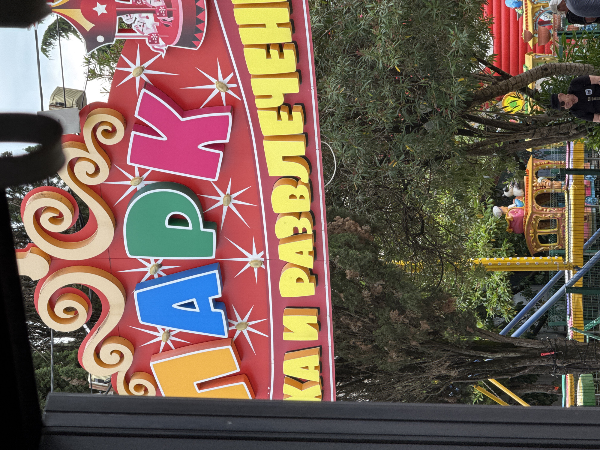 This media file captures a vibrant, colorful sign advertising a "парк развлечений" (amusement park) in Sirius, Russia. The sign features large, bold, three-dimensional letters in shades of pink, green, blue, and yellow, set against a bright red background adorned with white starbursts. Decorative golden swirls are also part of the sign's design.
In the background, glimpses of an amusement park can be seen. These include parts of rides with bright red, yellow, and blue structures, as well as lush green trees and foliage, suggesting an outdoor setting. The lighting appears to be natural daylight, possibly overcast or late afternoon, as there are no strong shadows.
A few people are visible in the background, including one person wearing a black cap and shirt, partially obscured by a tree. Another individual is seen wearing a light-colored cap and a darker shirt. The overall impression is of a fun and lively atmosphere typically associated with amusement parks.
The visible text on the sign clearly reads "ПАРК РАЗВЛЕЧЕНИЙ," which translates to "Amusement Park." The image seems to be taken from a slightly low angle, possibly from a vehicle, given the dark frame at the bottom of the image.