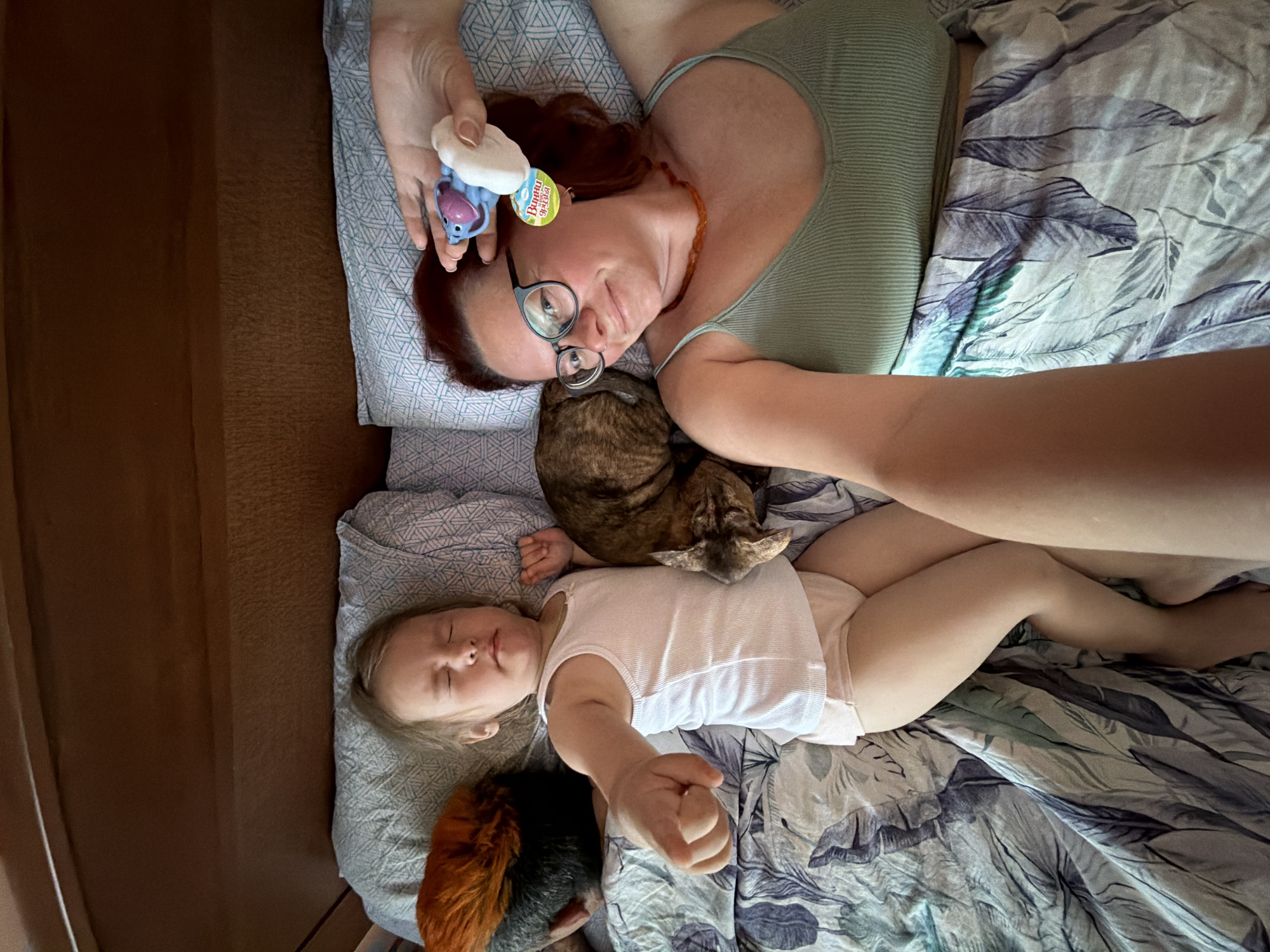 The image captures an intimate overhead view of a woman, a baby, a cat, and a chicken resting together on a bed indoors. The scene suggests a moment of domestic tranquility, possibly a nap or relaxed playtime.
The woman, with reddish-brown hair and wearing glasses, lies on her back on a patterned pillow. She is dressed in a ribbed light green top and has an amber-colored necklace. Her expression is calm, with a slight, gentle gaze towards the camera. She holds a blue and white toy, possibly a squeaky toy, in her right hand. Visible text on the toy's tag is in Cyrillic, reading "ВИННИ ЗВЕРЯТА" (VINNI ZVERYATA), translating to "Winnie Animals."
To her left, a tabby cat with striped fur is curled up, resting its head near the woman's shoulder and the baby's head. The baby, appearing to be a toddler, lies peacefully on their back, seemingly asleep or resting, wearing a white sleeveless bodysuit and light-colored briefs. Their eyes are closed, and hair is light blonde.
At the foot of the bed, near the baby's feet, a chicken or rooster with vibrant orange and black feathers is also visible, resting. The bedding consists of a light-colored pillow with a geometric pattern and a duvet featuring a leafy, watercolor-like design in blues and grays. The overall lighting suggests daytime indoors.
The image conveys a tender and affectionate interaction between family members, including pets, in a home setting, located in Chaikovsky, Russia.