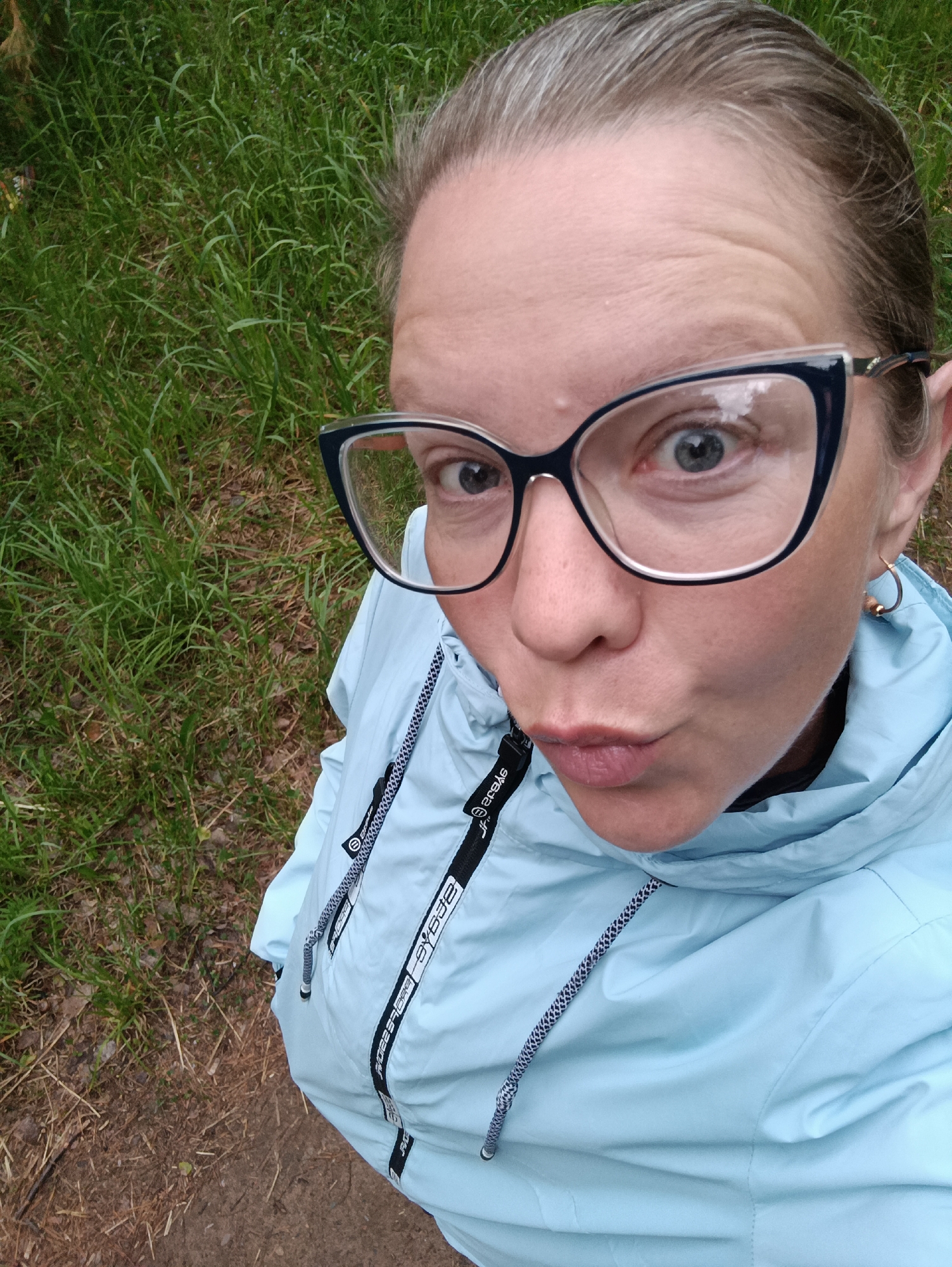 A woman with graying hair pulled back, wearing black-framed glasses and a light blue jacket, is taking a selfie outdoors. She is puckering her lips towards the camera. She has a single gold hoop earring visible. She appears to be standing on a dirt or grassy surface. In the background is green grass, some of which is growing in a patchy area of bare earth, possibly in a park or wooded area. There is no visible indication that this image was taken in the city of Chaikovsky, Russia.